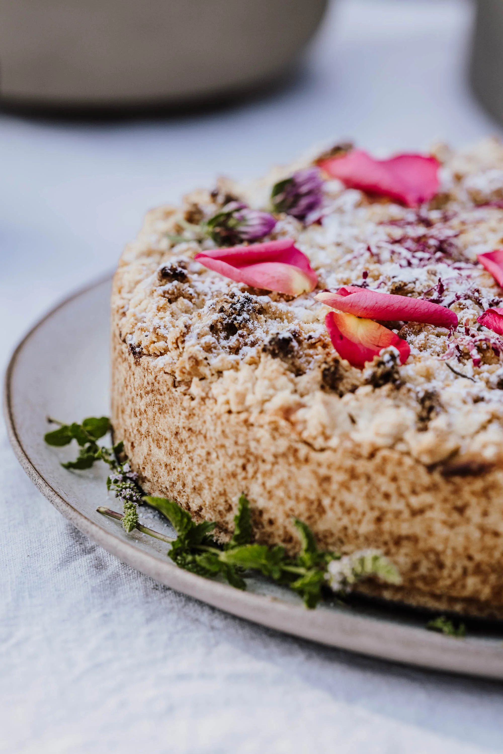 Ein runder Kuchen mit Vanille- oder Sahne-Füllung, bedeckt mit Streuseln, rosa Rosenblättern und kleinen Blumen, auf einem weißen Teller