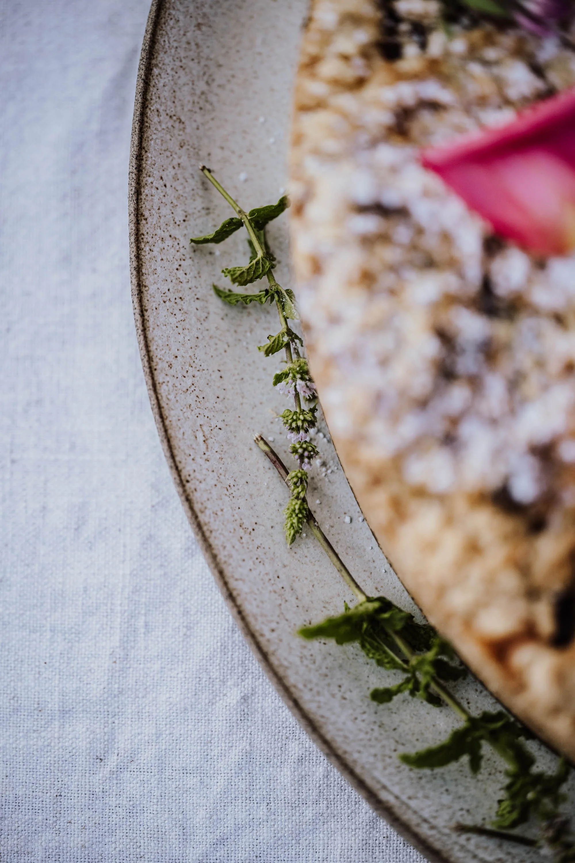 Nahaufnahme eines Tellers mit einer Scheibe Kuchen oder Törtchen, dekoriert mit frischen Kräutern, auf einem weißen Stofftisch.