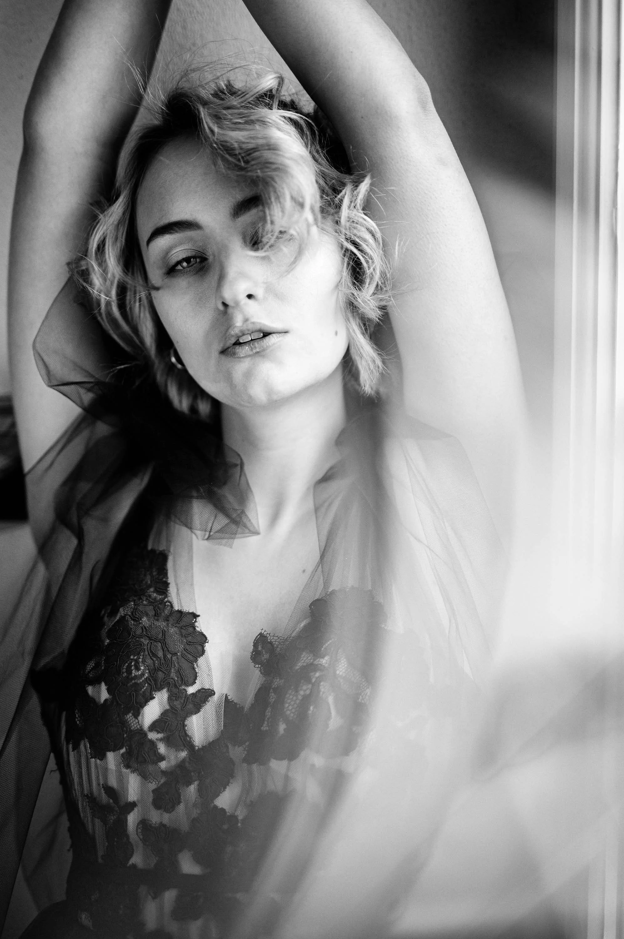 Schwarz-Weiß-Fotografie eines jungen Mädchens mit lockigem Haar, das leicht mit einem Auge geschlossen schaut und in einem schwarzen Spitzenkleid und transparenter Tülljacke gekleidet ist, mit einem Arm über dem Kopf, vor einem Fensterstehend.