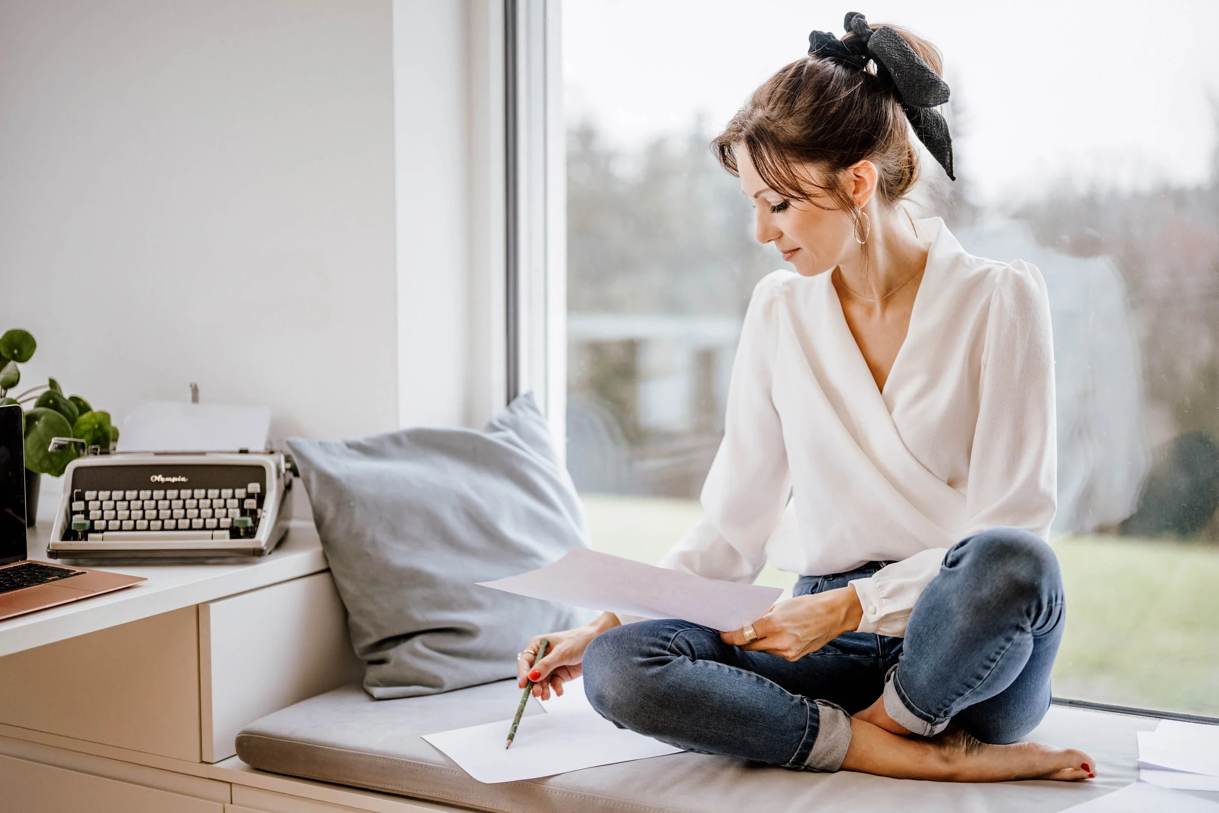 Eine Frau sitzt am Fenster auf einem Sitz mit Kissen, liest Papier und schreibt mit einem Bleistift, während sie entspannt und konzentriert aussieht.