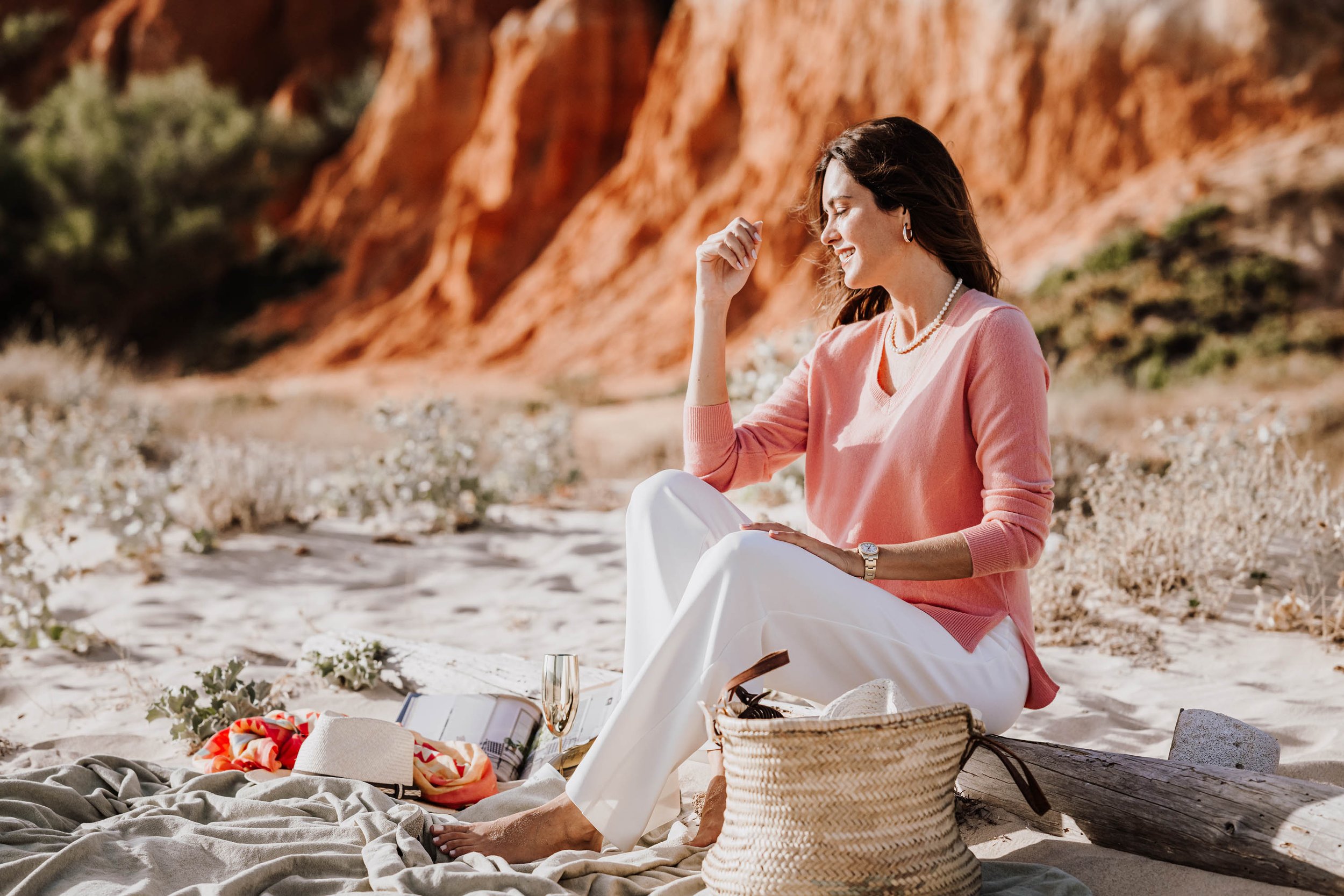Eine lachende Frau sitzt am Strand mit Blick auf rote Felsen im Hintergrund, umgeben von Picknickutensilien und einem Korb.