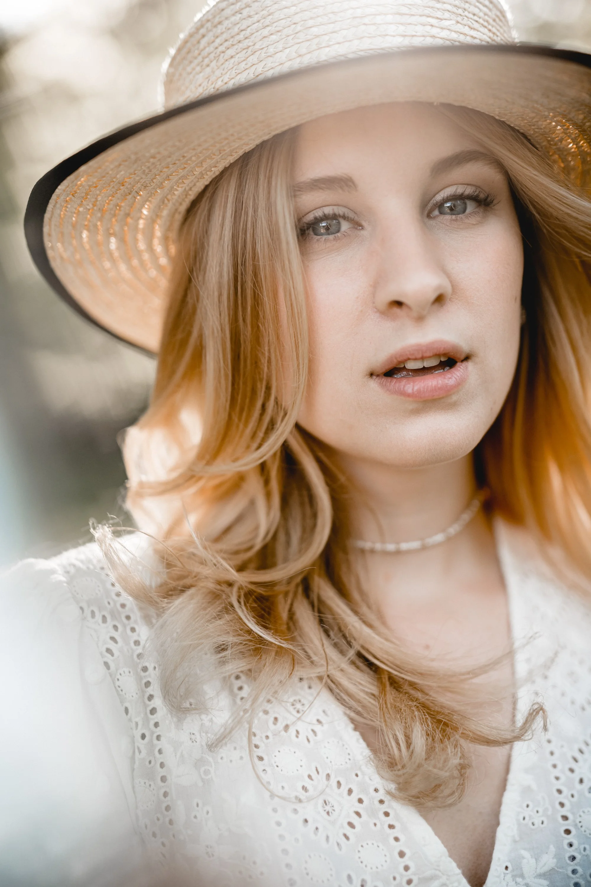 Junge Frau mit locker fallen gelassenen, blonden Haaren, trägt einen Strohhut mit schwarzem Band und eine weiße Bluse mit Stickerei, im Freien bei Sonnenlicht.