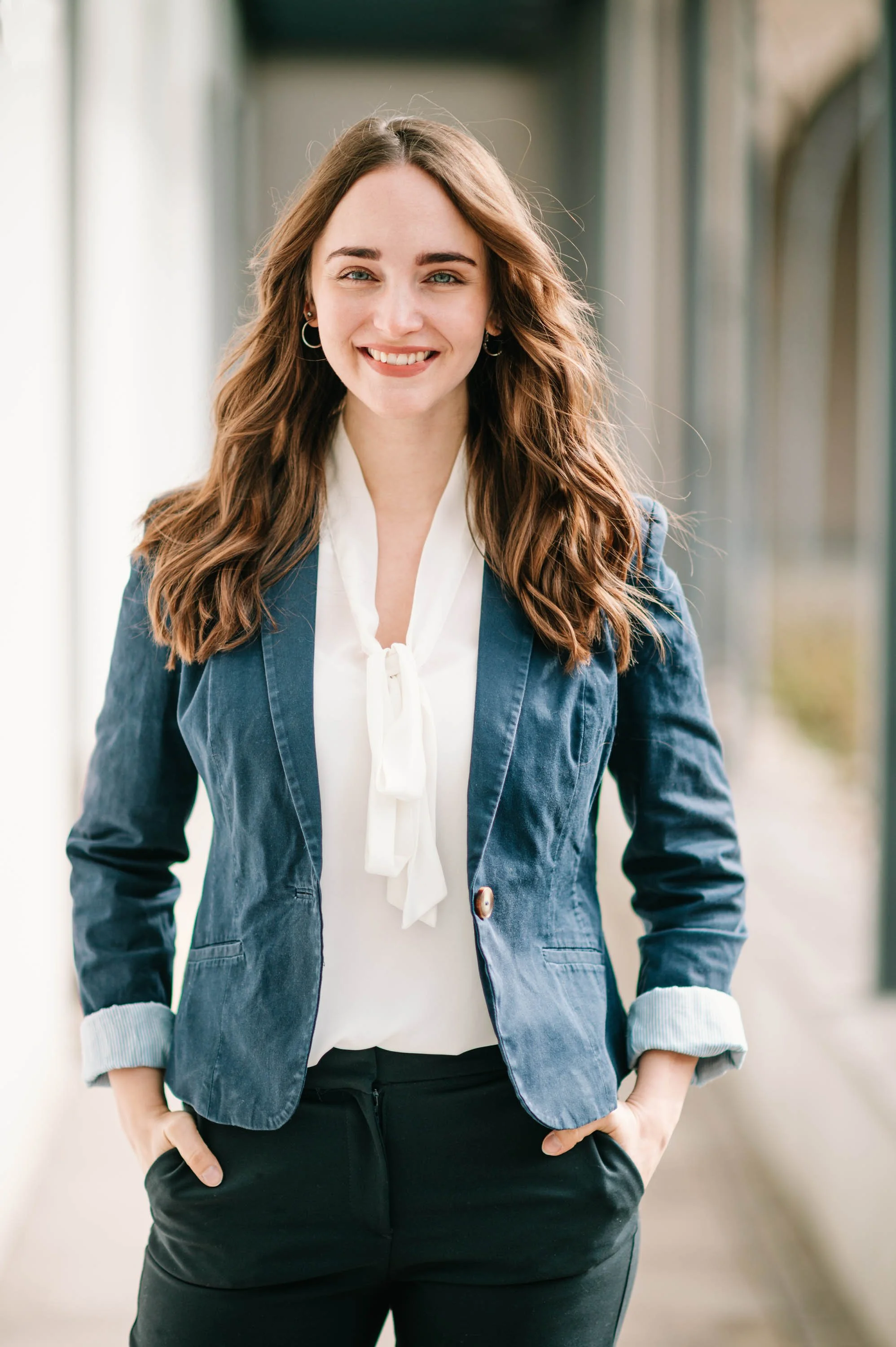 Lächelnde junge Frau mit lang, lockigem braunem Haar trägt eine weiße Bluse, einen blauen Blazer und schwarze Hosen. Sie steht draußen an einem sonnigen Tag, mit einer Hand in der Tasche und sieht direkt in die Kamera.