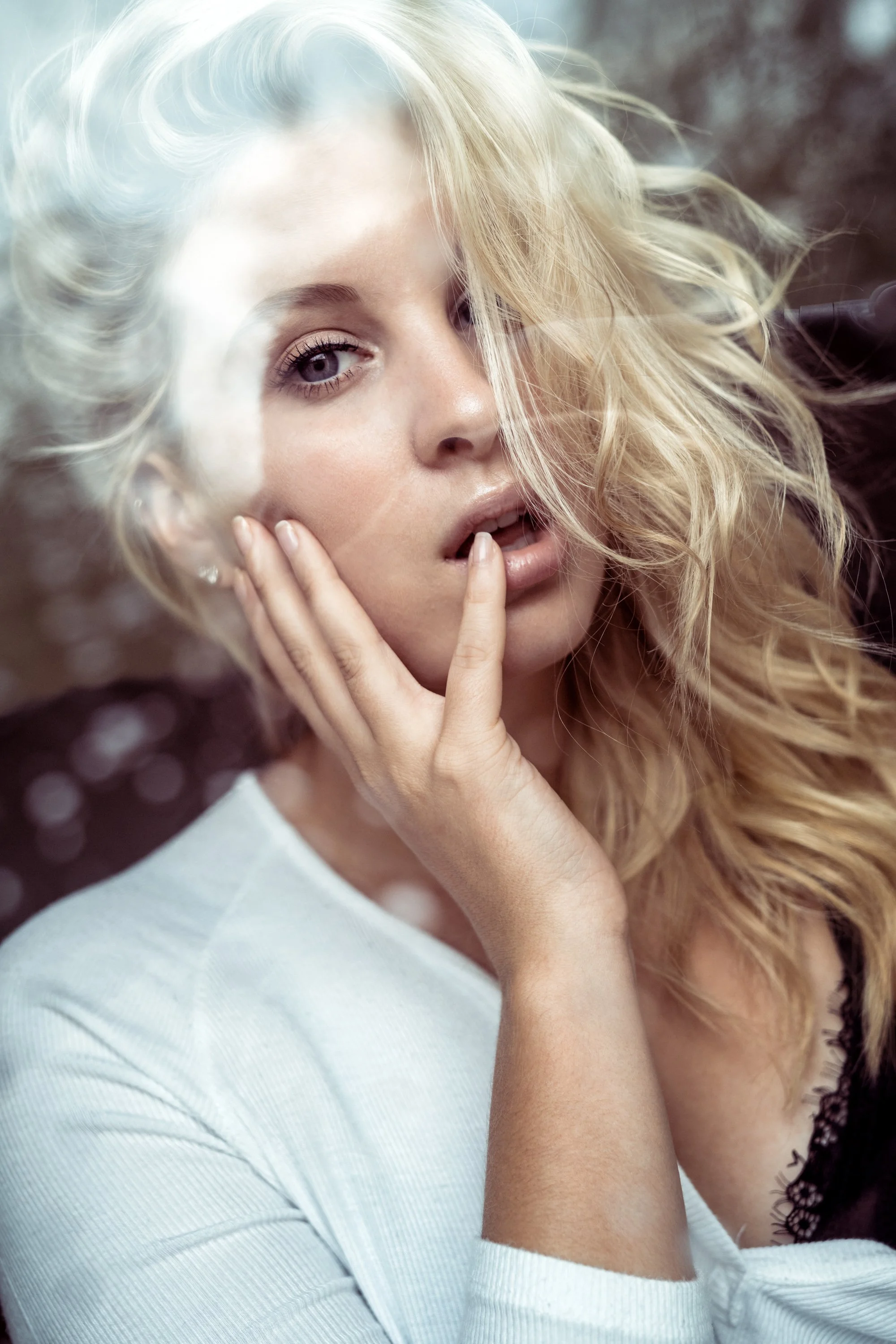 Ein junges blondes Model mit lockigem Haar, nah aufgenommen, mit den Händen im Gesicht und leicht geöffnetem Mund, vor unscharfem Hintergrund mit Lichtereffekten.