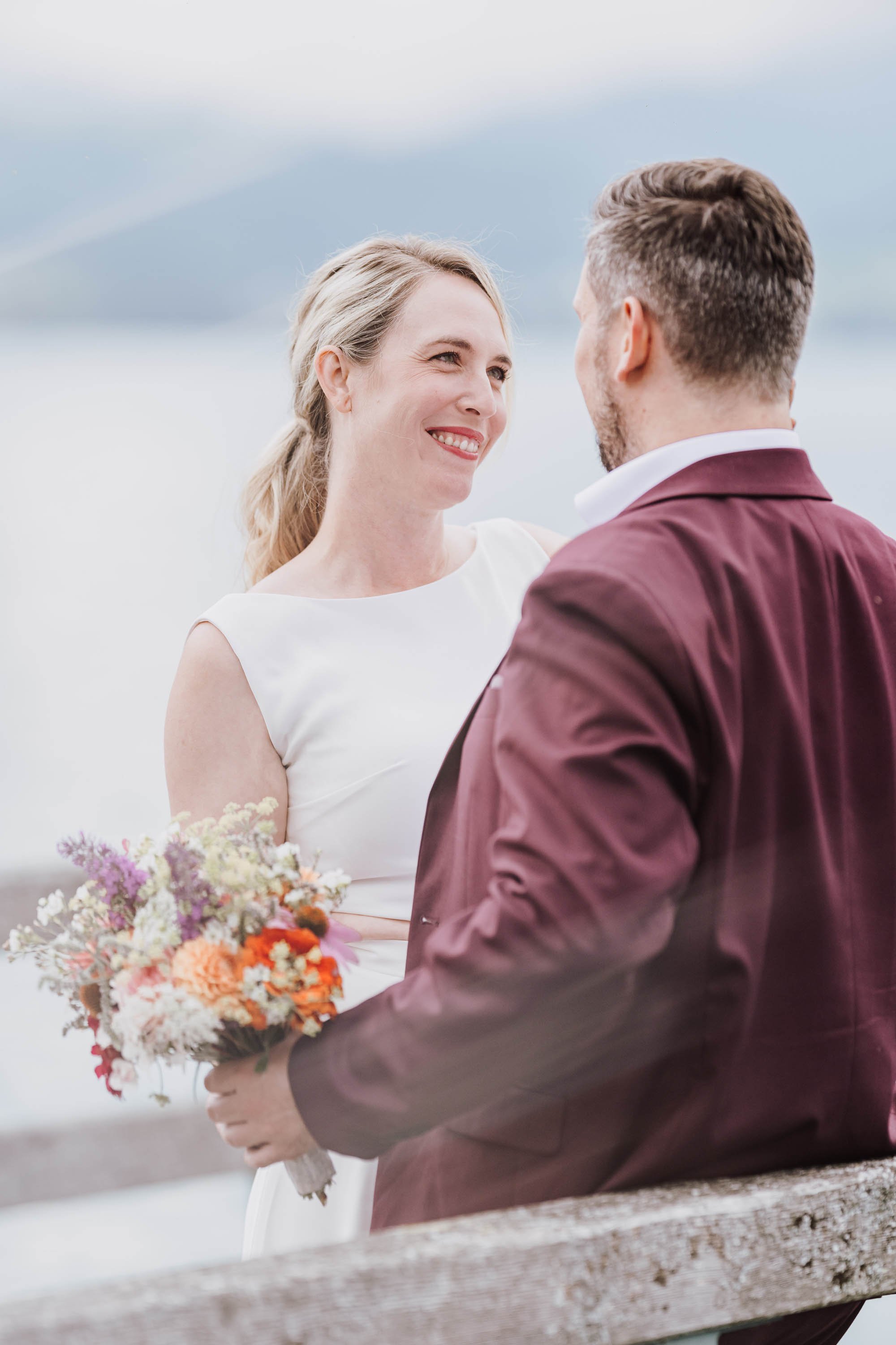 Ein Brautpaar steht am Wasser, die Braut hält einen bunten Blumenstrauß und lächelt den Bräutigam an, während er ihm in die Augen schaut.