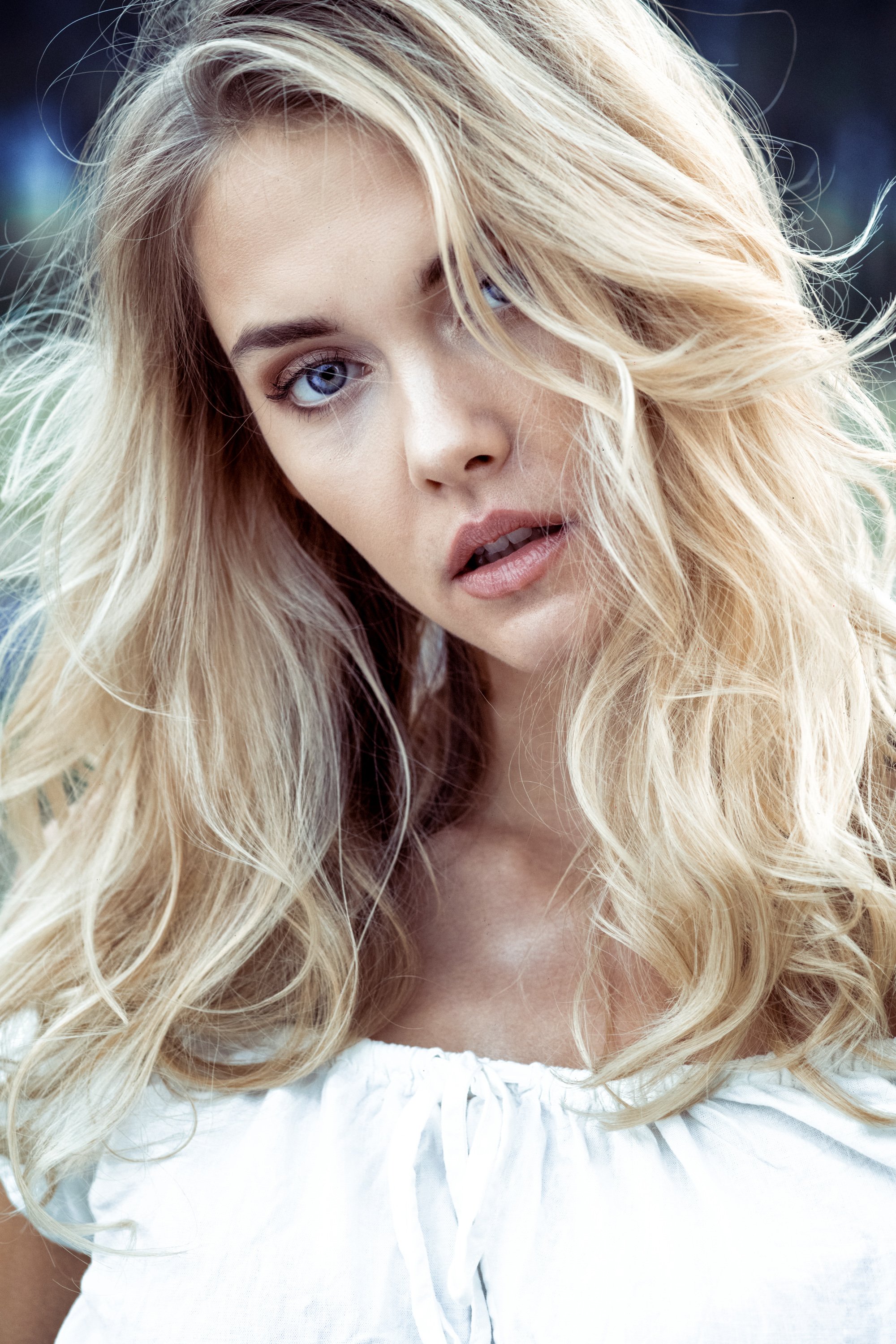 Nahaufnahme einer blonden Frau mit welligem Haar, blauen Augen, leicht geöffneten Lippen, trägt ein weißes Kleid im Freien.