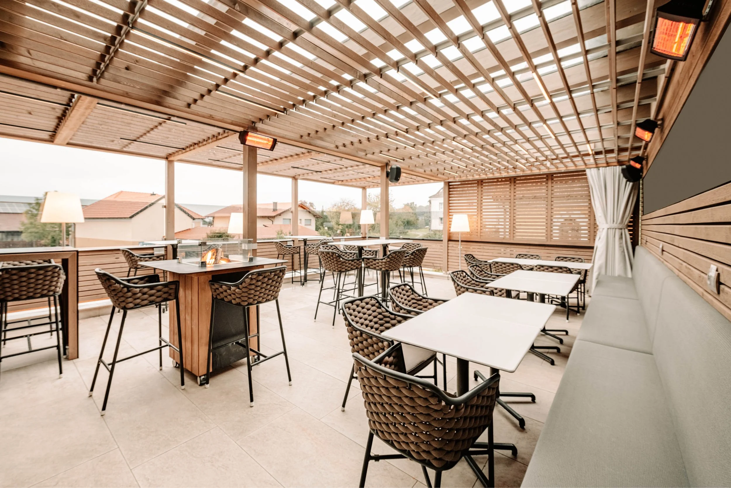 Gemütlicher, halbexterner Sitzbereich mit Holzwänden und -Decke, mehreren Tischen und Stühlen, Dekor mit Lampen, Heizstrahlern und einer Lounge.