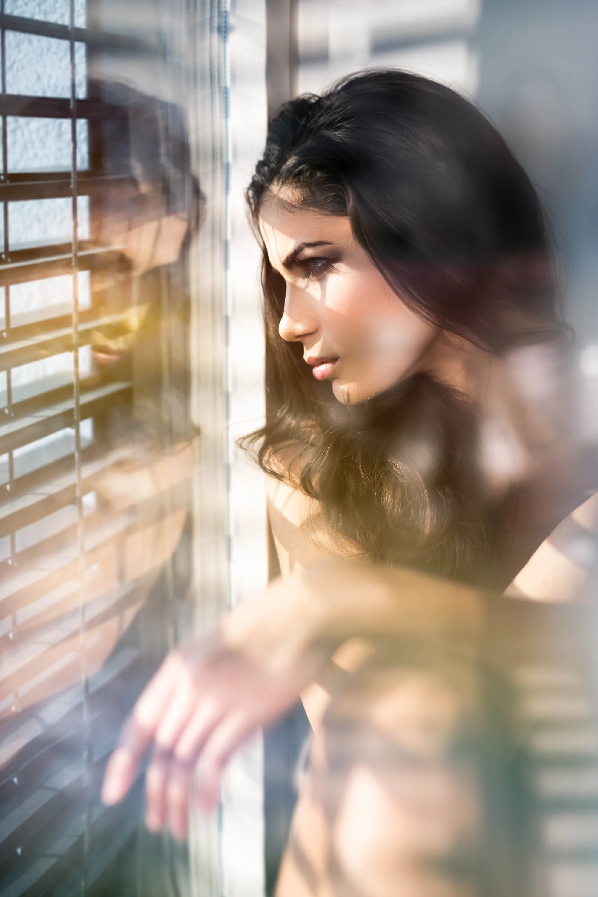 Junge Frau blickt durch eine Fensterjalousie nach draußen, Sonnenlicht fällt auf ihr Gesicht.