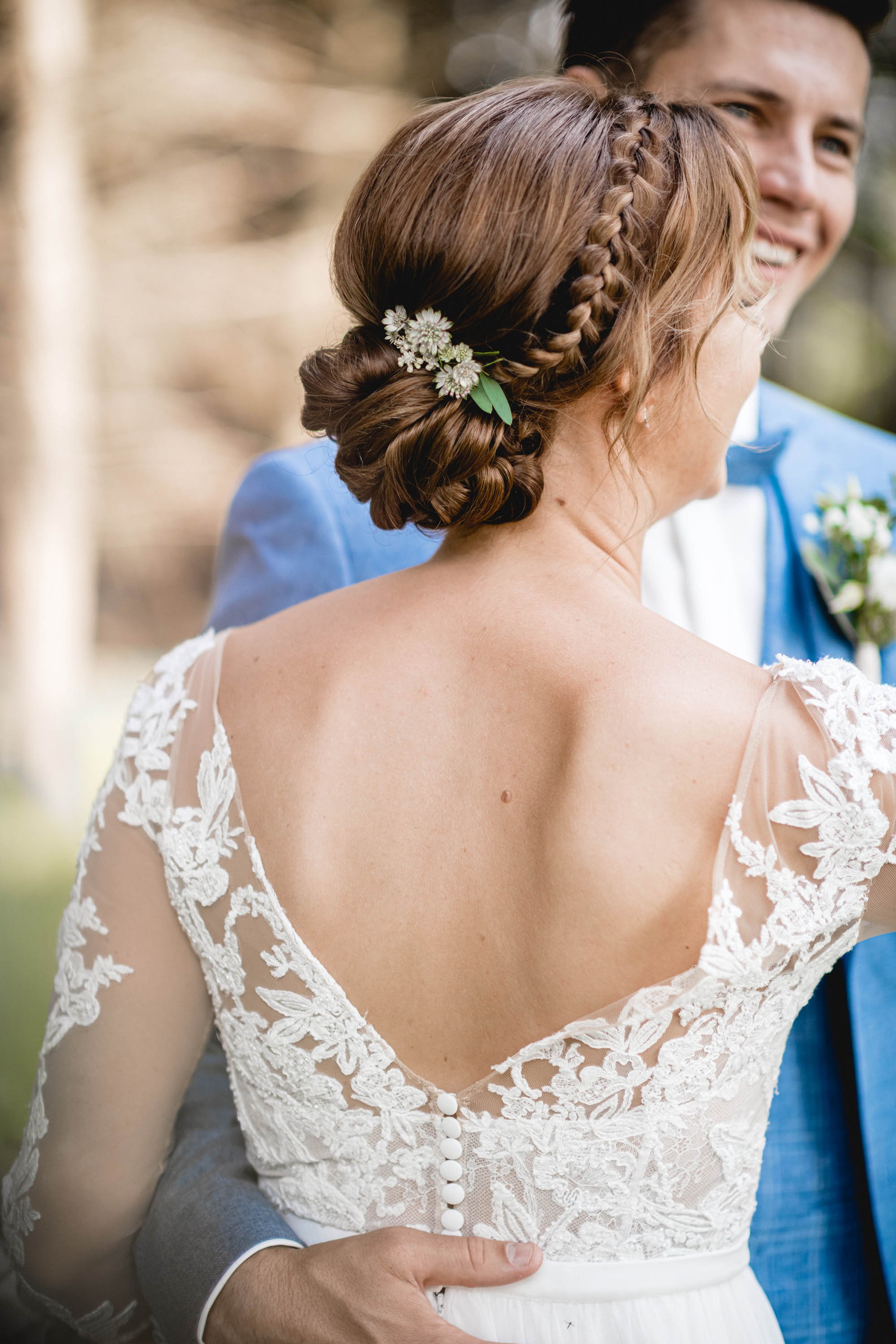 Nahe Aufnahmen eines Brautpaares bei ihrer Hochzeitszeremonie im Freien, Fokus auf die Braut mit schönem Brautkleid und geflochtenem Haarknoten mit Blumen, während der Bräutigam im blauen Anzug lächelt.