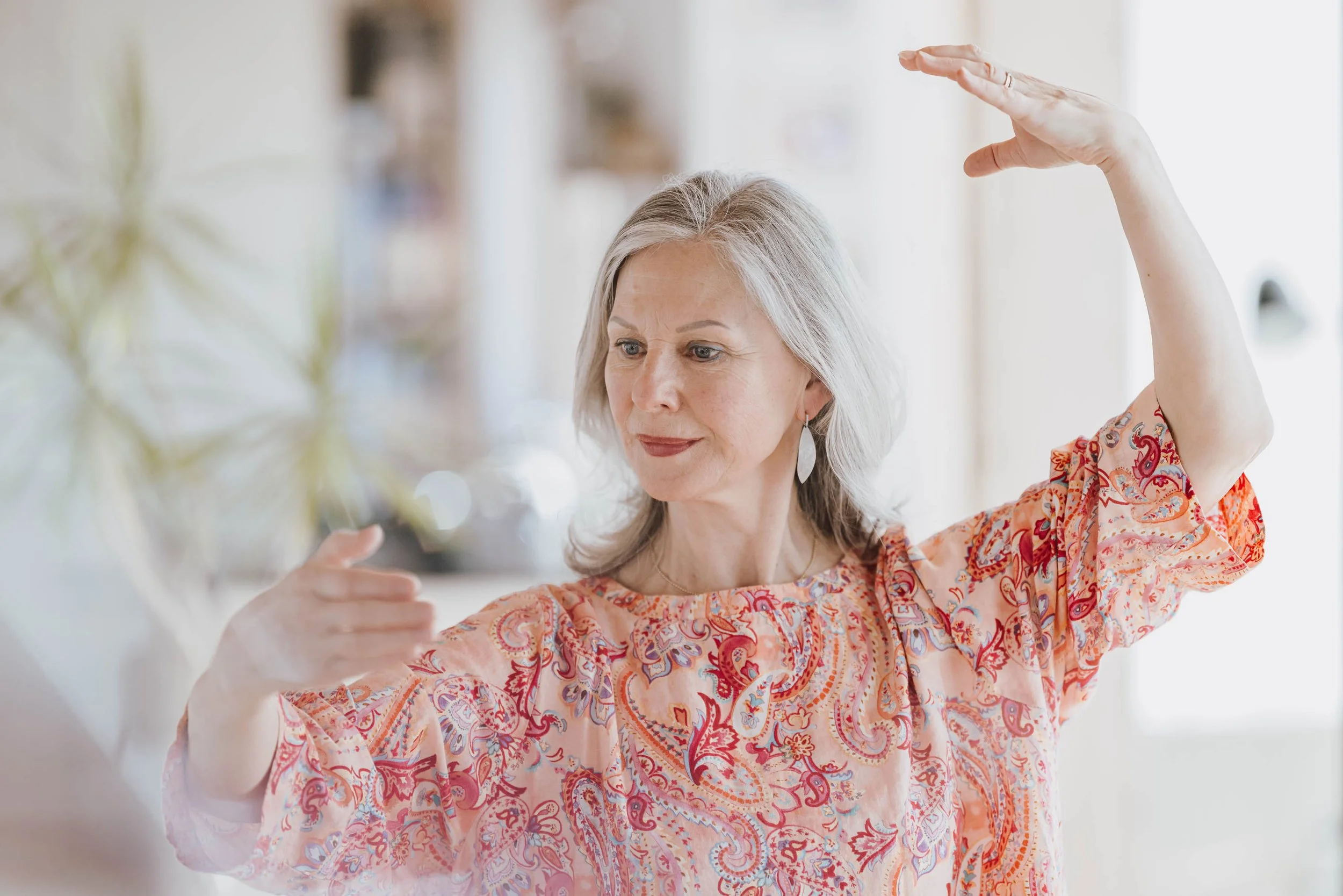 Ältere Frau mit grauem Haar, die im Wohnzimmer Yoga macht, trägt einen bunten, paisley-musterhaften Overall.