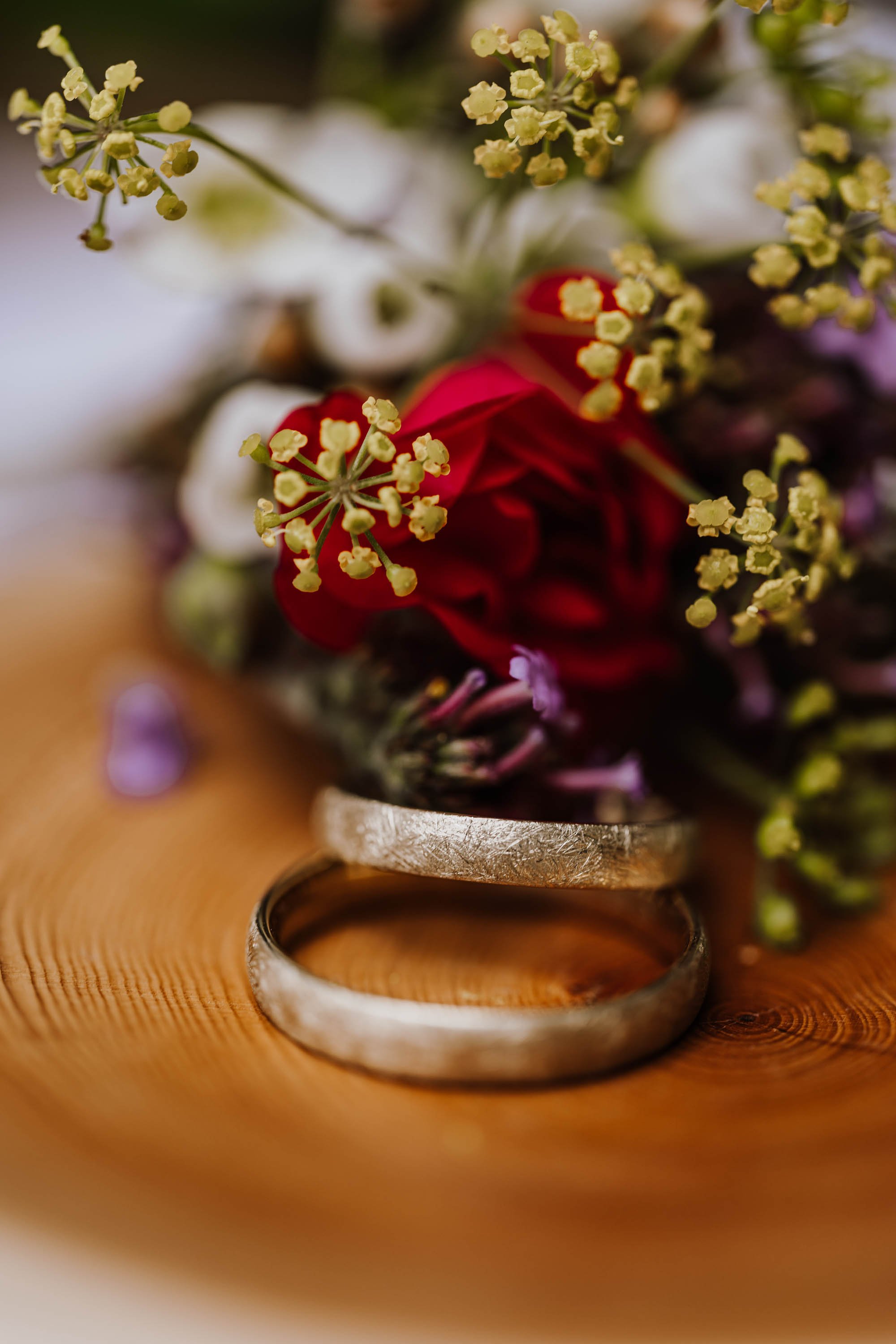 Zwei silberne Eheringe liegen auf einem Holzuntergrund, umgeben von bunten Blumen und Blüten.
