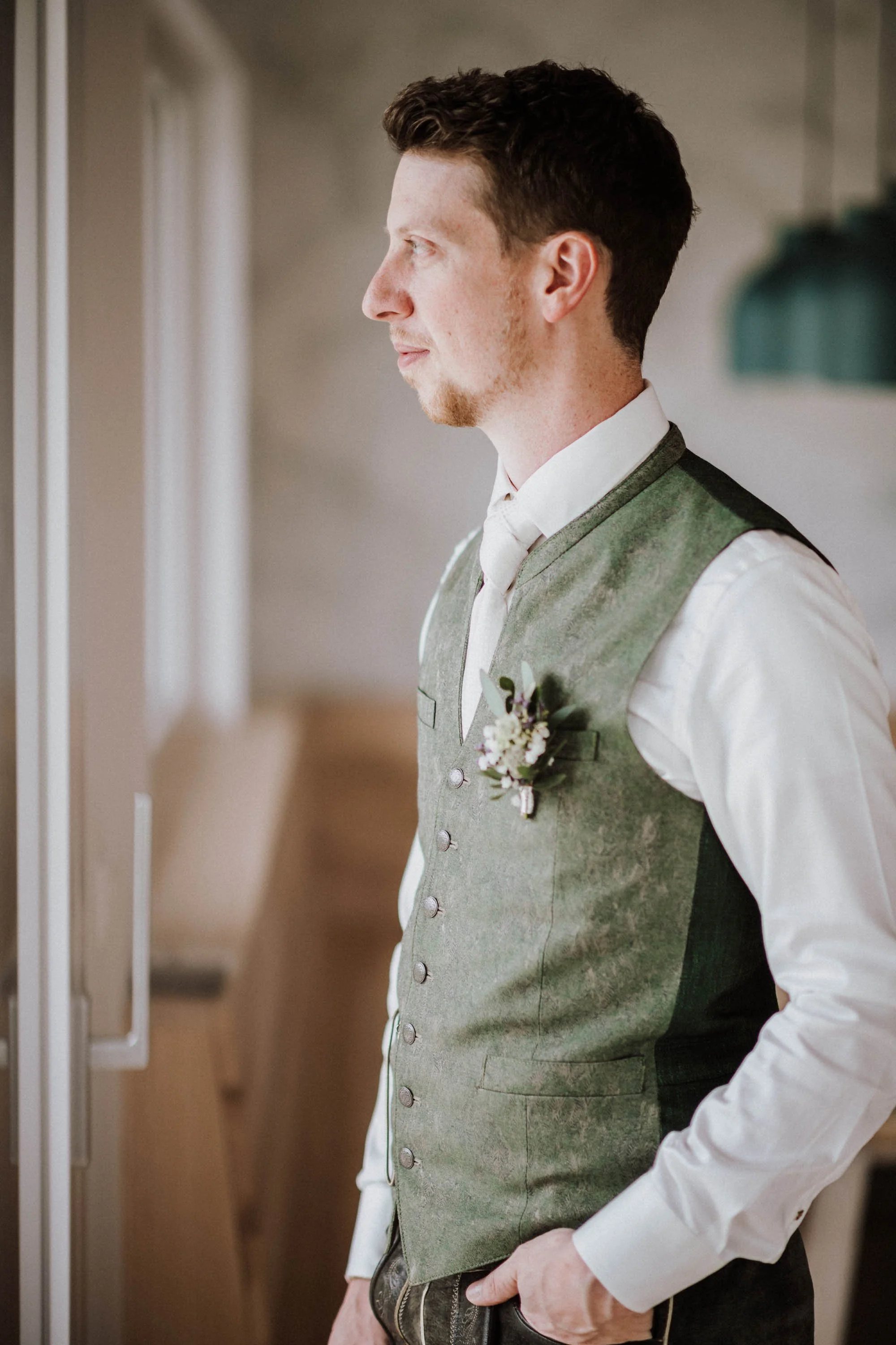 Ein junger Mann im traditionellen deutschen Dirndl-Look, steht seitlich, mit den Händen in den Taschen, vor einem Fenster, trägt eine weiße Hemd, eine grüne Weste mit Knöpfen und einen Blumenanstecker. Wohl bei einer besonderen Gelegenheit.