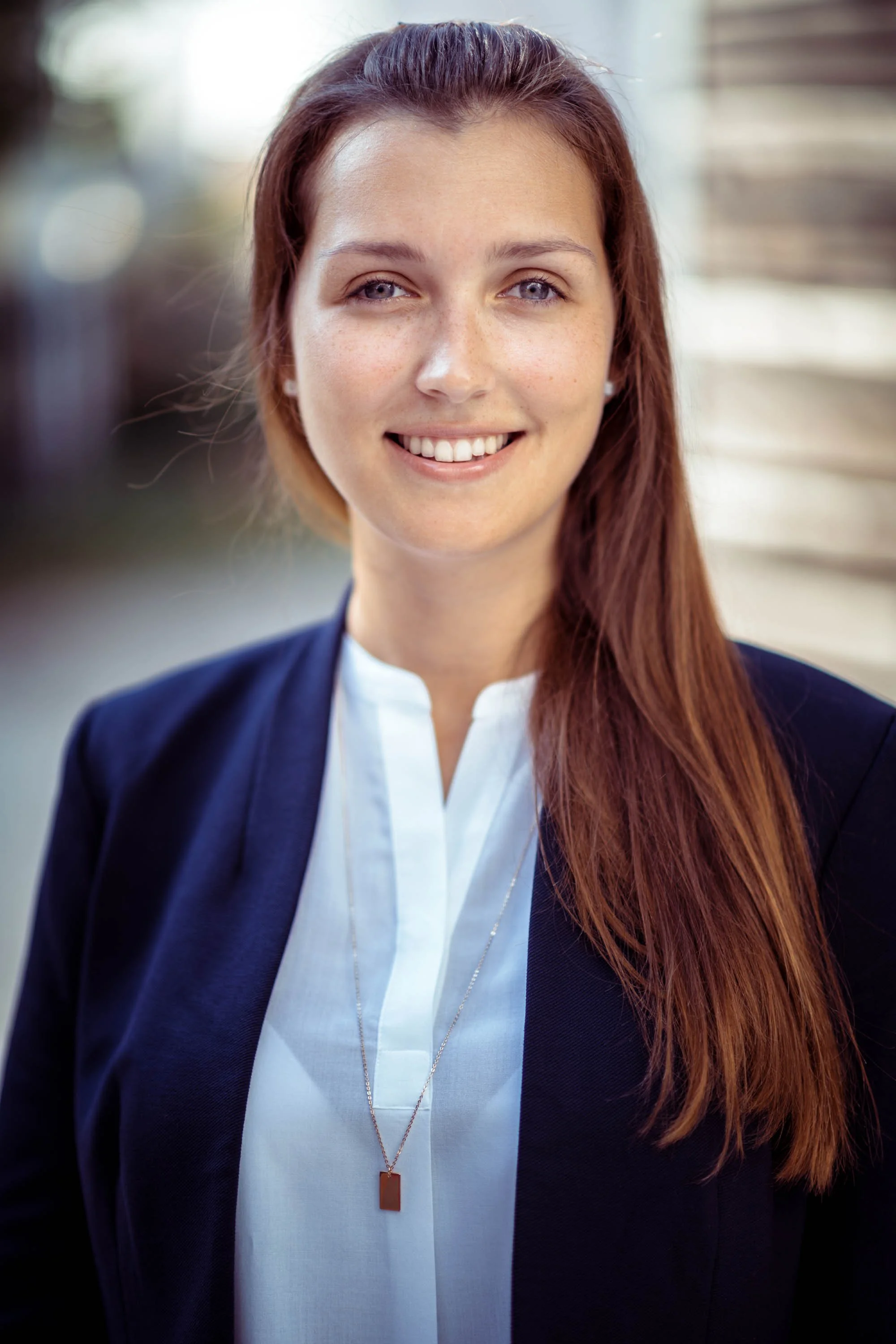 Porträt einer lachenden jungen Frau mit roten Haaren, die eine weiße Bluse und eine dunkle Jacke trägt, vor einem urbanen Hintergrund.