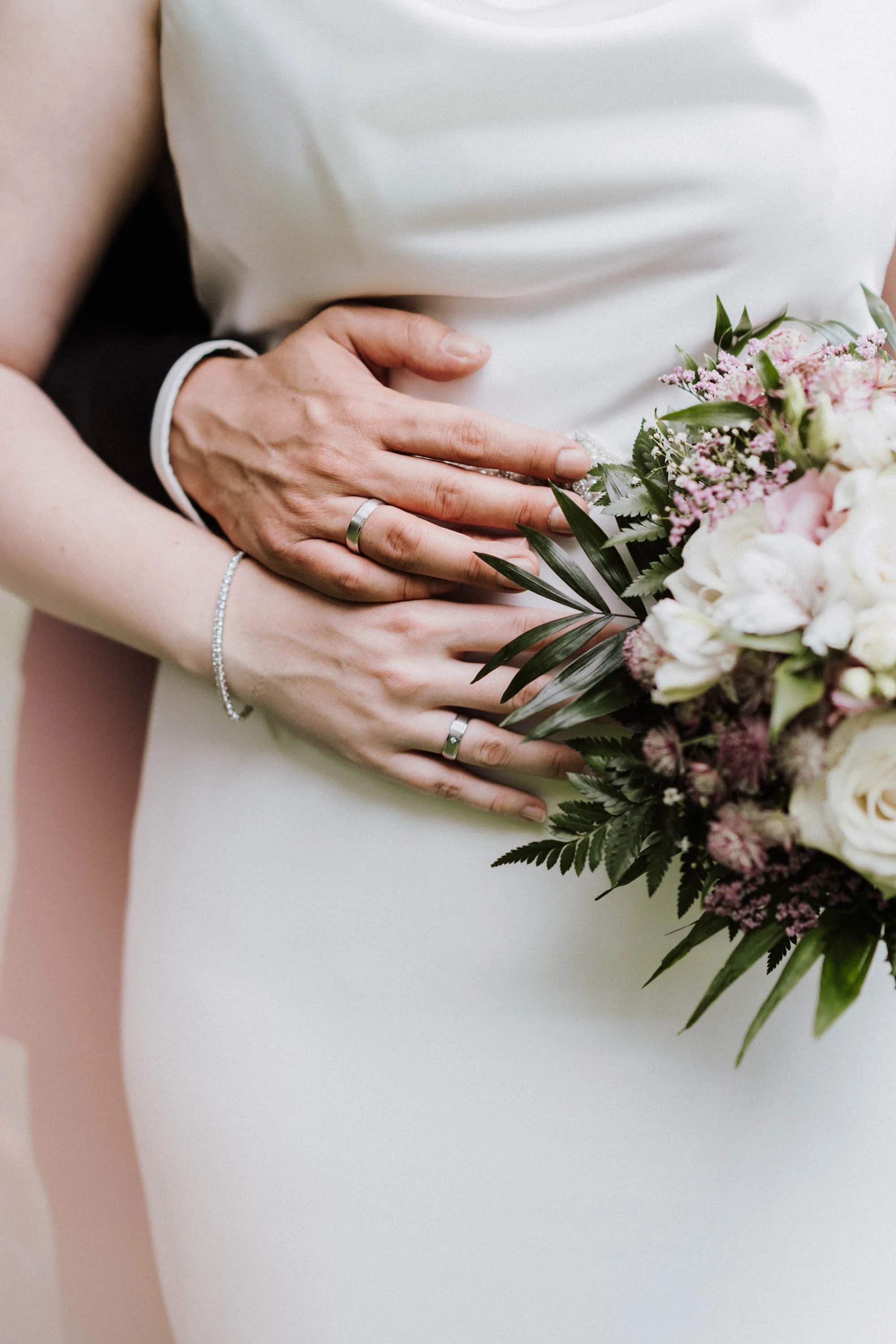 Nahaufnahme der Hände eines Brautpaares mit Eheringen, die eine Blumenstrauß halten.