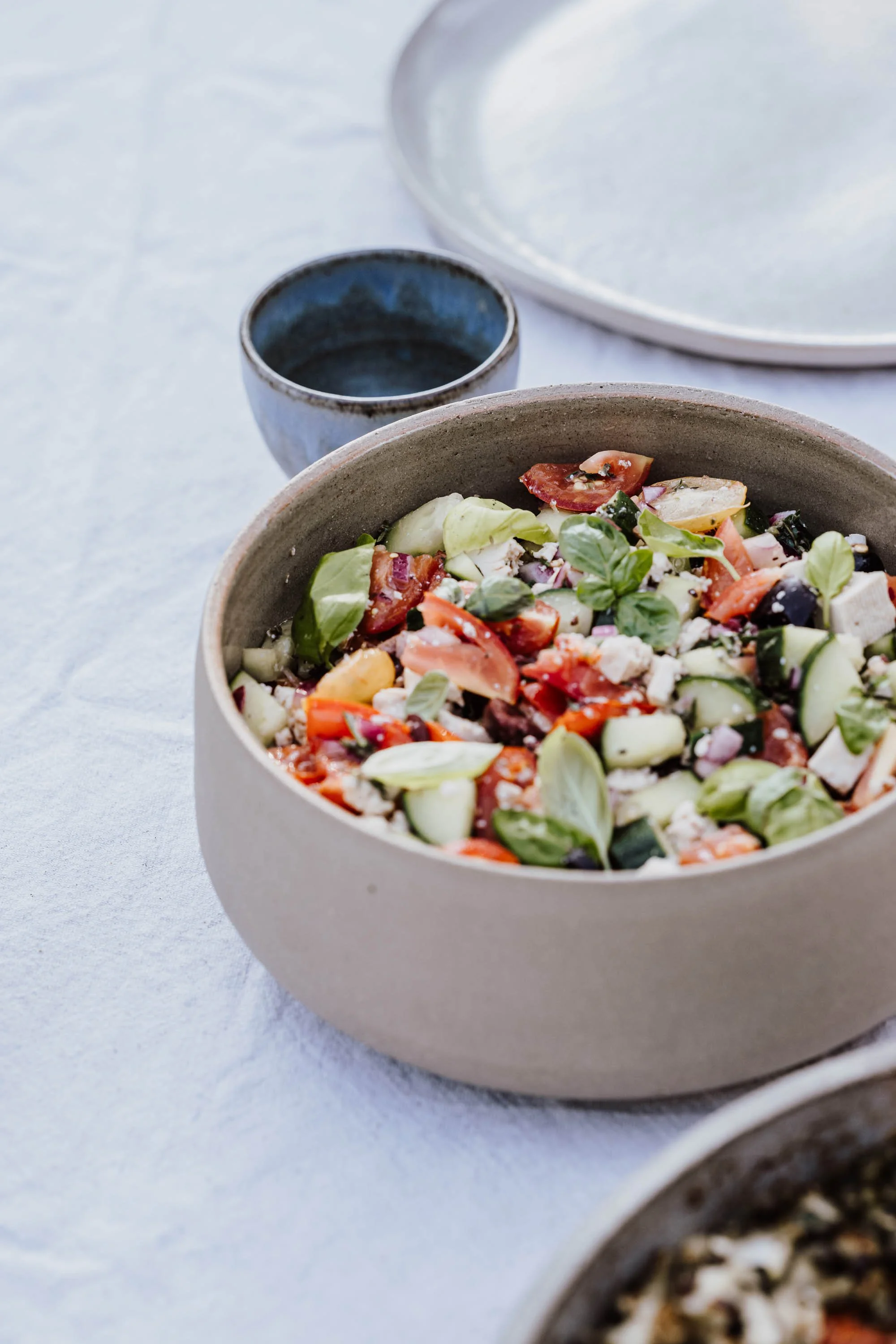 Eine Schüssel mit gemischtem Sommersalat auf einem weißen Tisch, im Hintergrund eine kleine blaue Schale und ein weißer Teller.