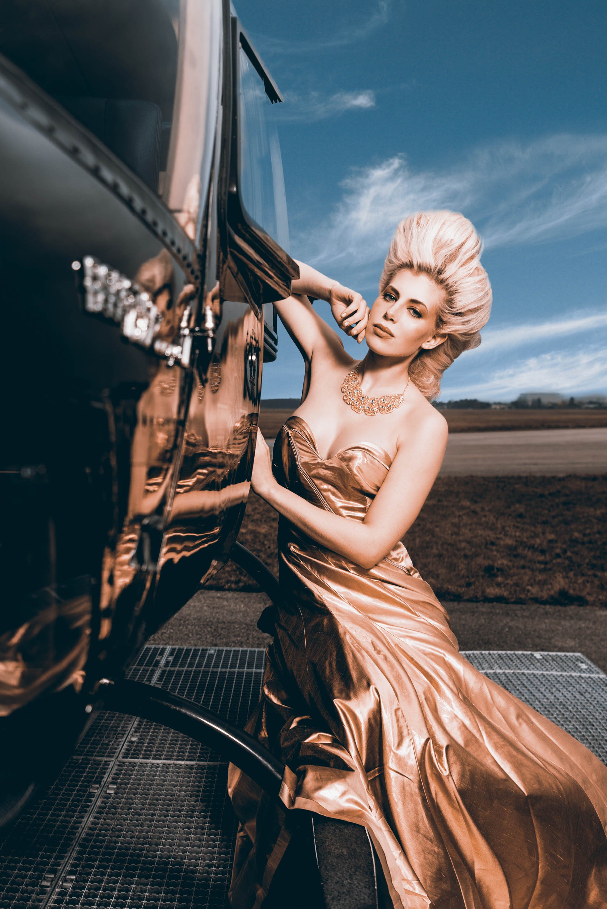 Eine Frau in einem eleganten, goldfarbenen Abendkleid mit Schmuck steht draußen vor einem schwarzen Auto, während sie die Kamera betrachtet.