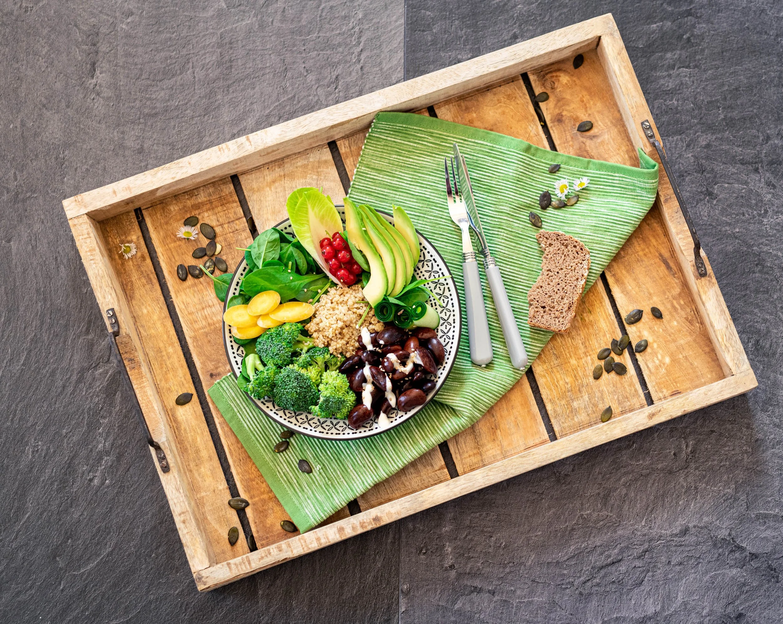 Eine Schüssel mit verschiedenem Bio-Gemüse und schwarzen Oliven auf einem grünen Tuch, auf einem Holztablett mit einer Gabel, einem Messer und einer Brotscheibe
