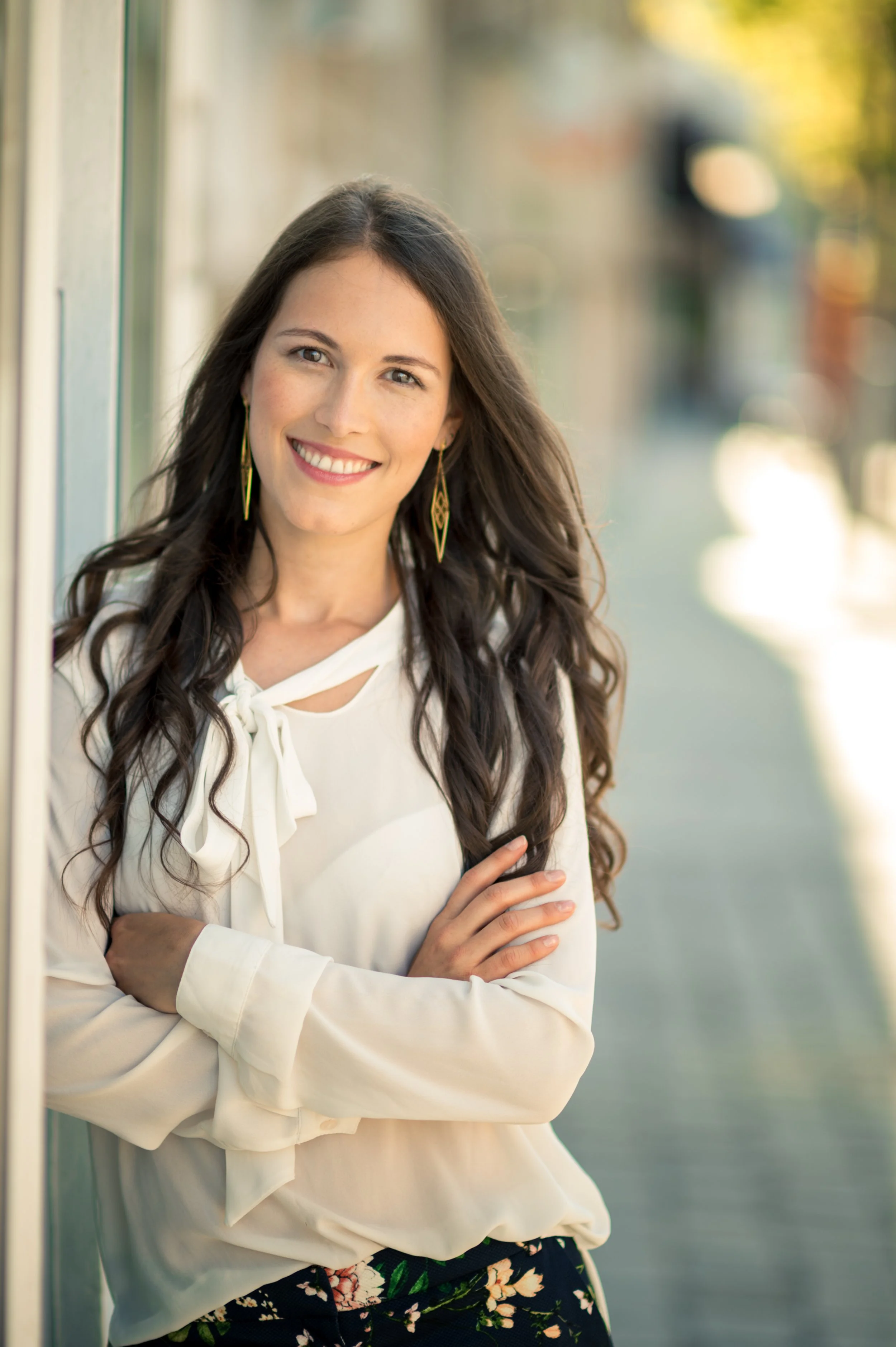 Junge Frau mit langen, dunklen, gewellten Haaren, lächelnd, steht an einer Wand im Freien, trägt eine weiße Bluse mit Schleife und eine schwarze Hose mit Blumenmuster, dies ist ein Portraitfoto.