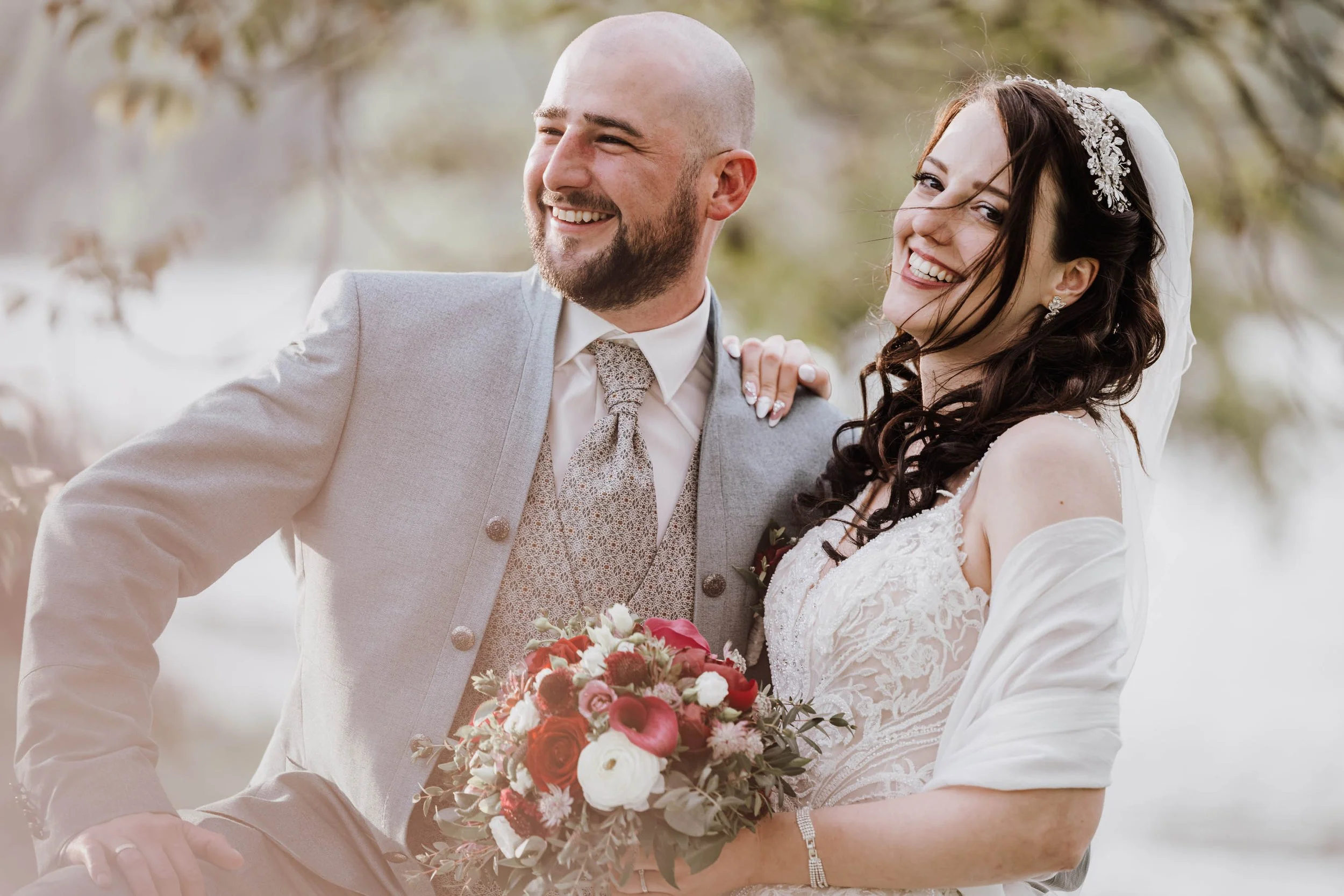 Ein glückliches Brautpaar bei einer Hochzeit im Freien, mit einer Frau in einem weißen Brautkleid und einem Mann in einem grauen Anzug, lächelnd und fröhlich, während sie einen Brautstrauß hält.