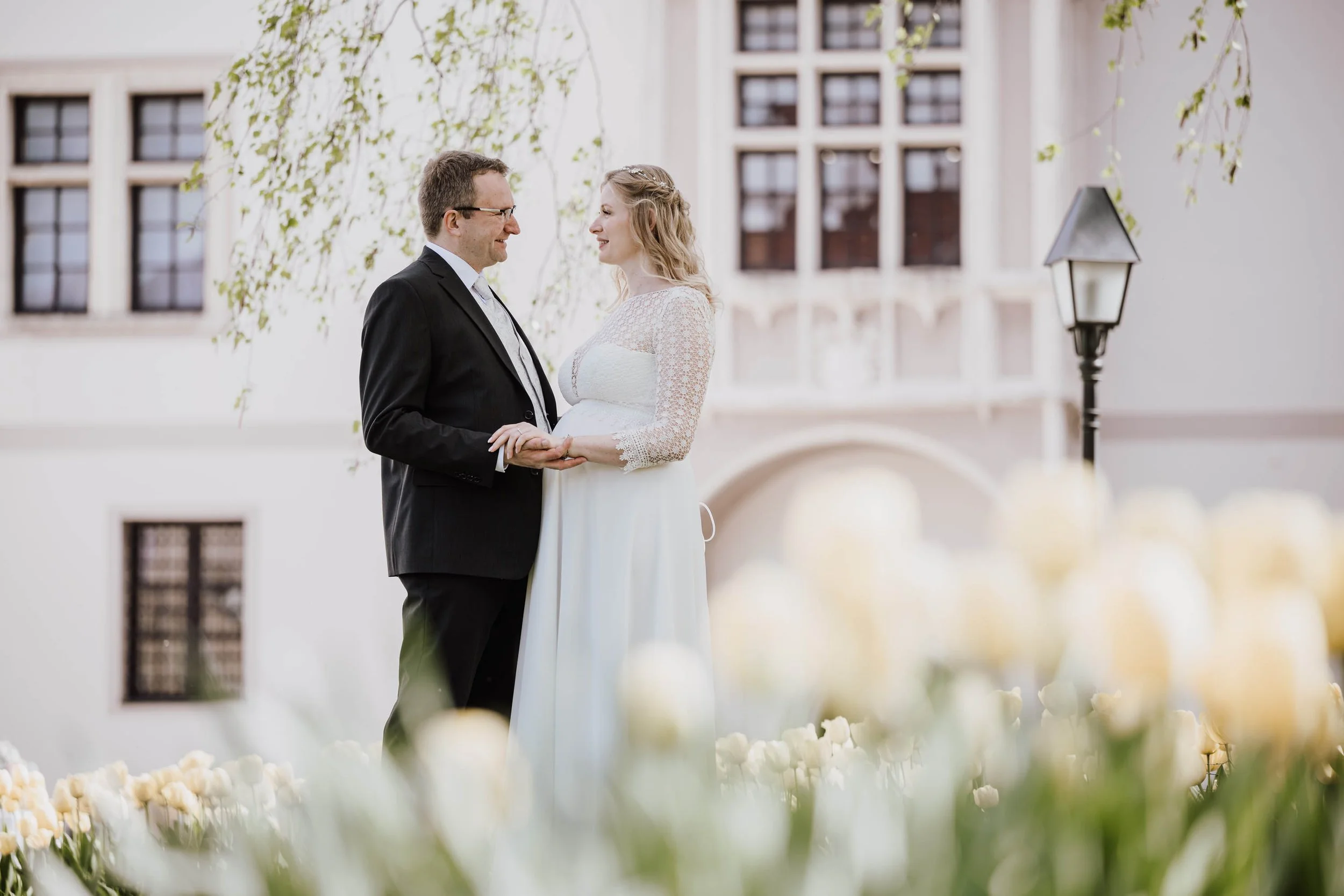 Ein Paar bei einer Hochzeitszeremonie im Garten, die Braut trägt ein weißes Kleid mit Spitze, der Bräutigam einen schwarzen Anzug. Im Hintergrund ein altes Gebäude und ein Straßenlaterne, im Vordergrund weiße Blumen.