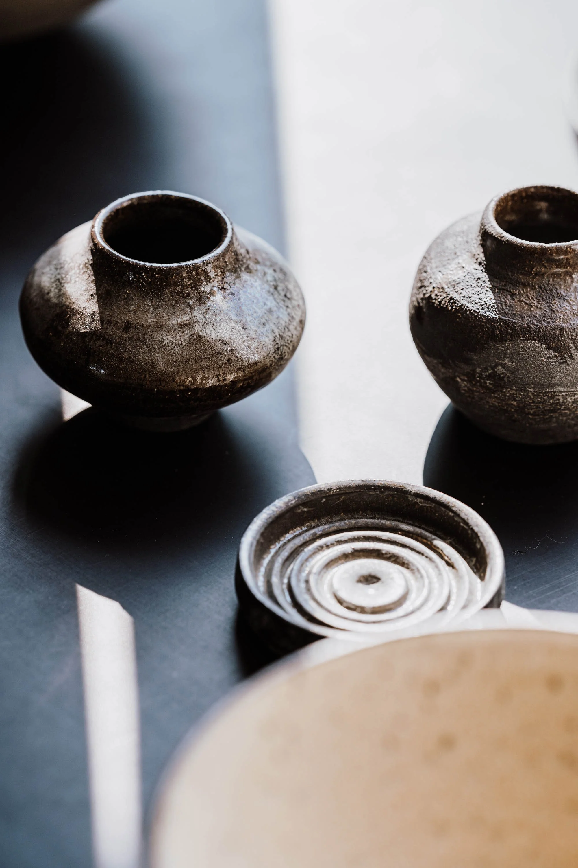 Zwei alte, braune Töpfergefäße und eine getöpferte Scheibe auf einer dunklen Oberfläche mit Licht- und Schatteneffekten.