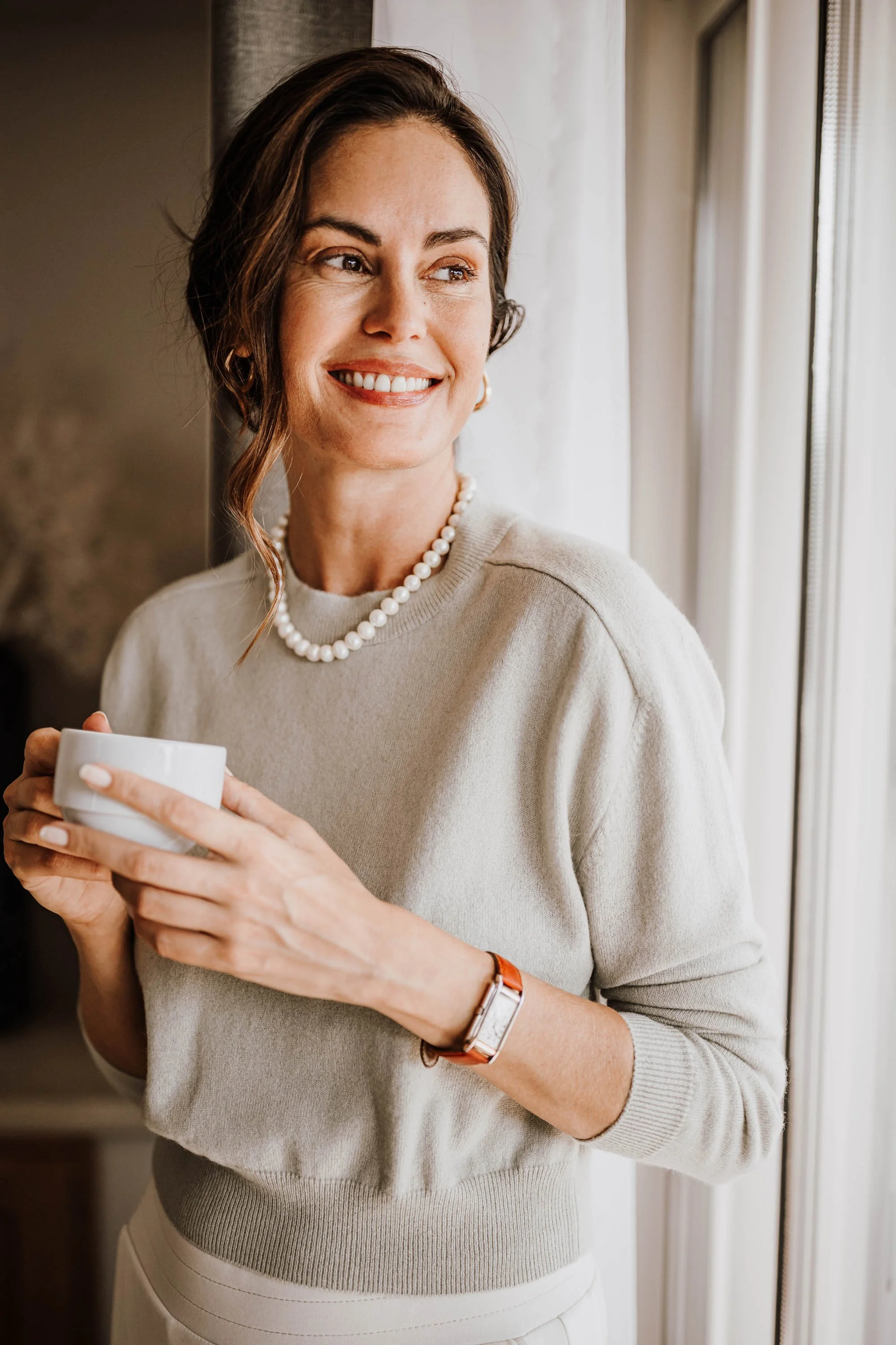 Eine Frau mit braunen, kurzen, gewellten Haaren, die eine Perlenkette trägt, steht neben einem Fenster, hält eine weiße Tasse und lächelt. Sie trägt eine beige Strickjacke, eine Uhr und einen Ringe-Ohrring. Das Bild vermittelt eine gemütliche und elegante Atmosphäre.