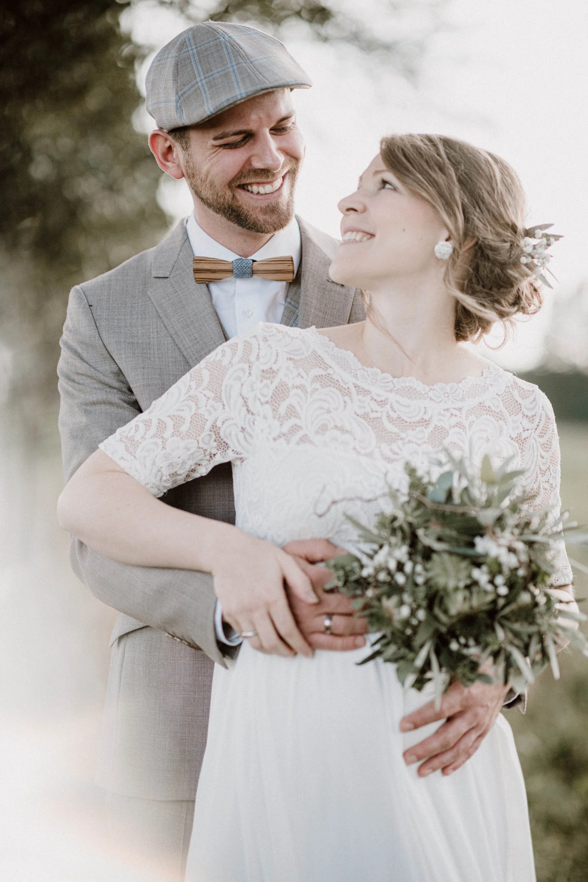 Ein glückliches Hochzeitspaar, das sich umarmt, draußen bei Sonnenuntergang, die Braut hält einen Blumenstrauß, beide lächeln sich an.