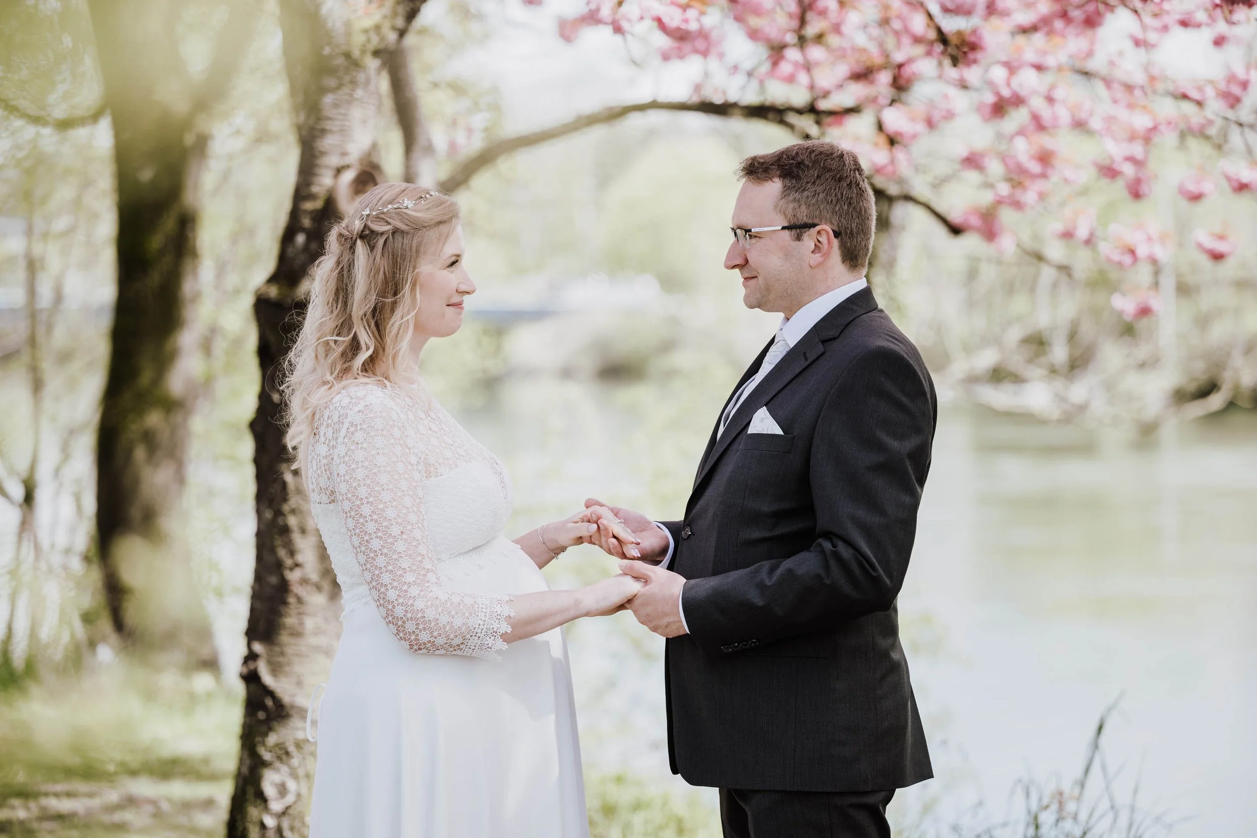 Ein Brautpaar steht sich unter blühenden Kirschbäumen gegenüber und hält Hände bei einer standesamtlichen Trauung im Freien. Die Braut trägt ein weißes Kleid mit Spitzenärmeln, der Bräutigam einen schwarzen Anzug und Brille.