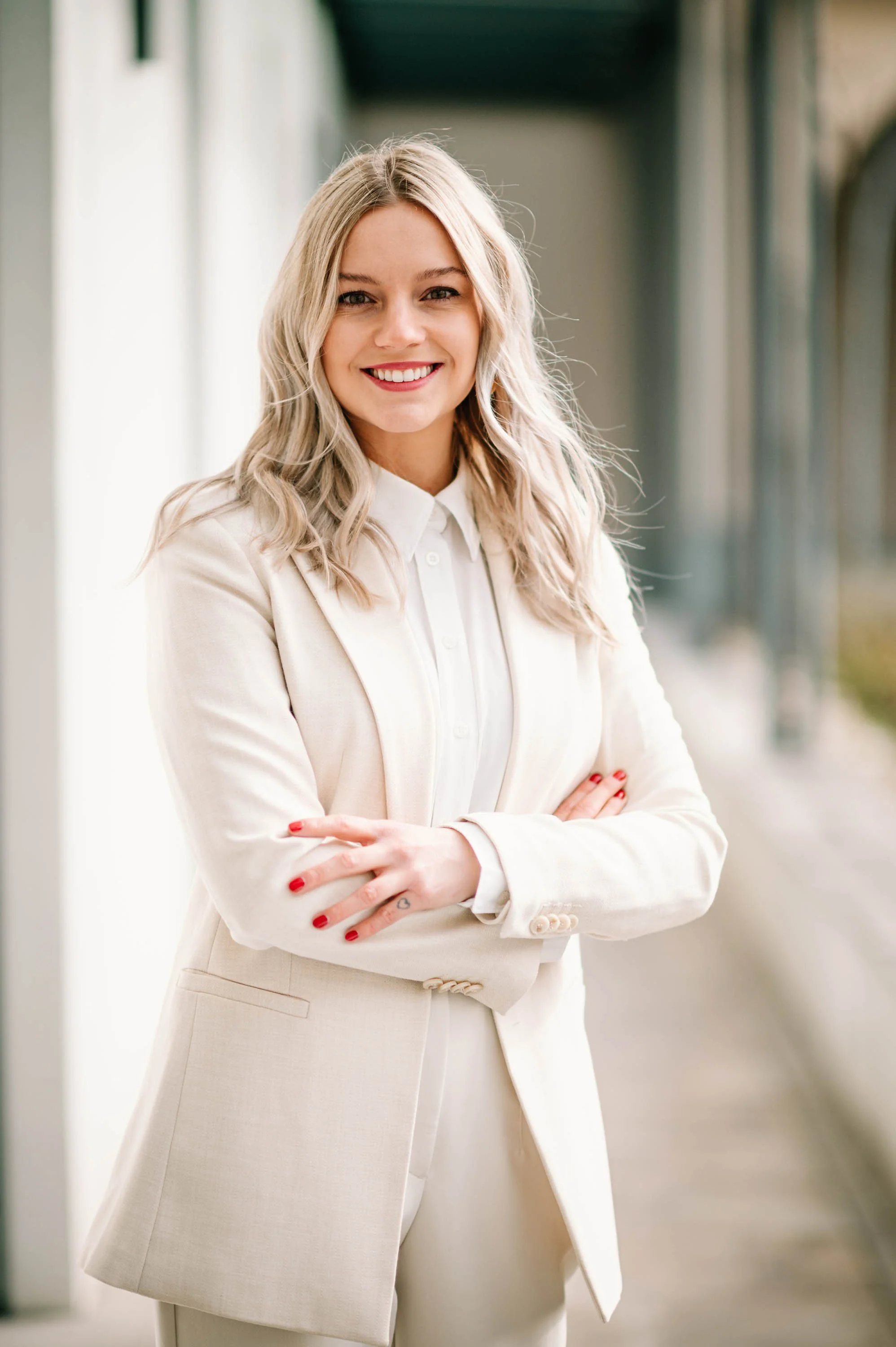 Eine lachende blonde Frau in einem weißen Anzug steht mit verschränkten Armen an einem Bahnsteig.
