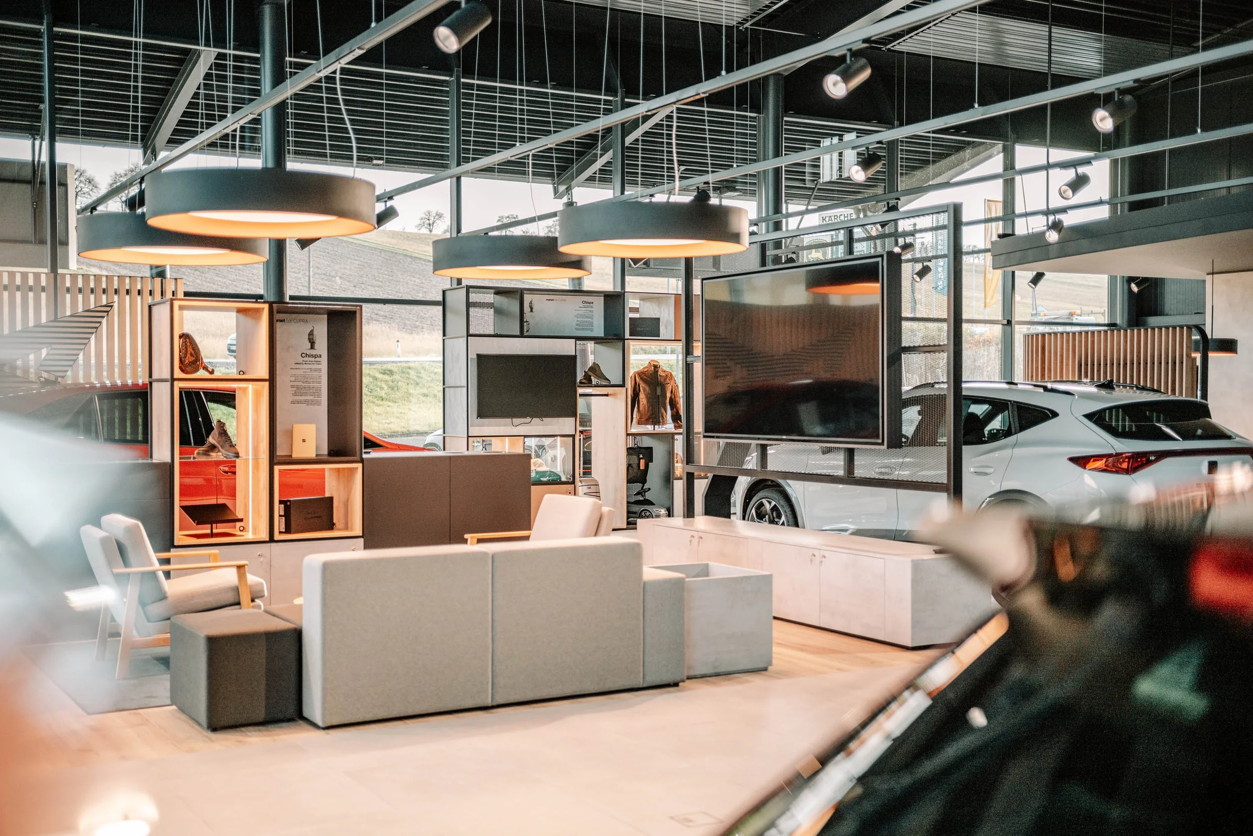 Moderne Autohalle mit ausgestellten Fahrzeugen, Sitzbereich mit Stühlen und Fernseher, große Fenster lassen natürliches Licht herein, dekorative Beleuchtung an der Decke.