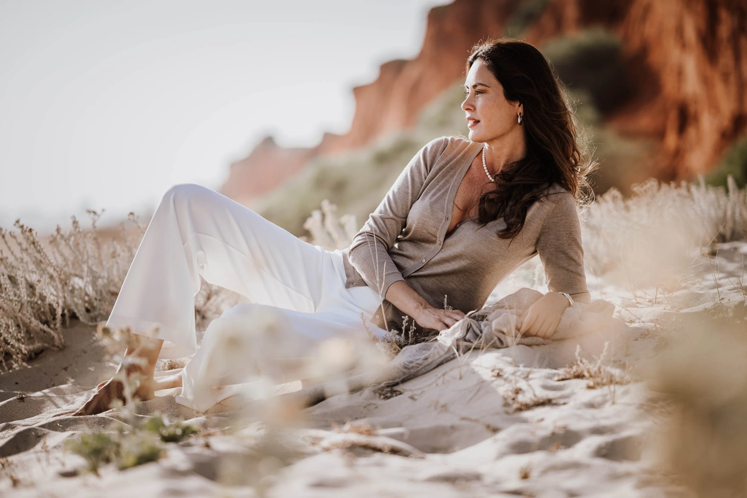 Eine Frau liegt entspannt am Strand zwischen Sand und Dünen, mit roten Klippen im Hintergrund, bei Sonnenlicht.