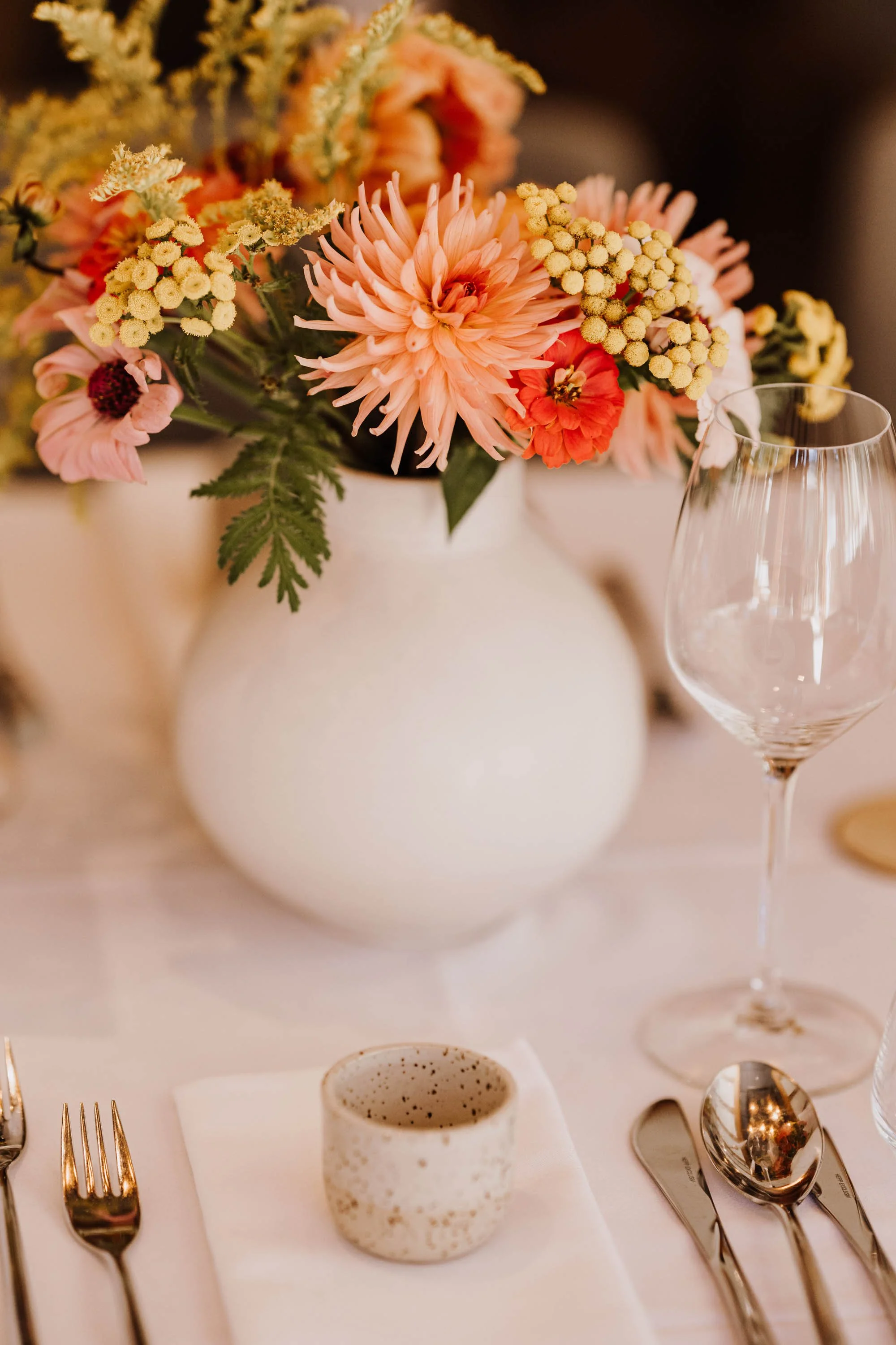 Blumenstrauß mit Pfirsichfarbigen und gelben Blumen in einer weißen Vase auf einem gedeckten Tisch, daneben leeres Weinglas, Messer, Gabel, Löffel und eine kleine Keramikschale.