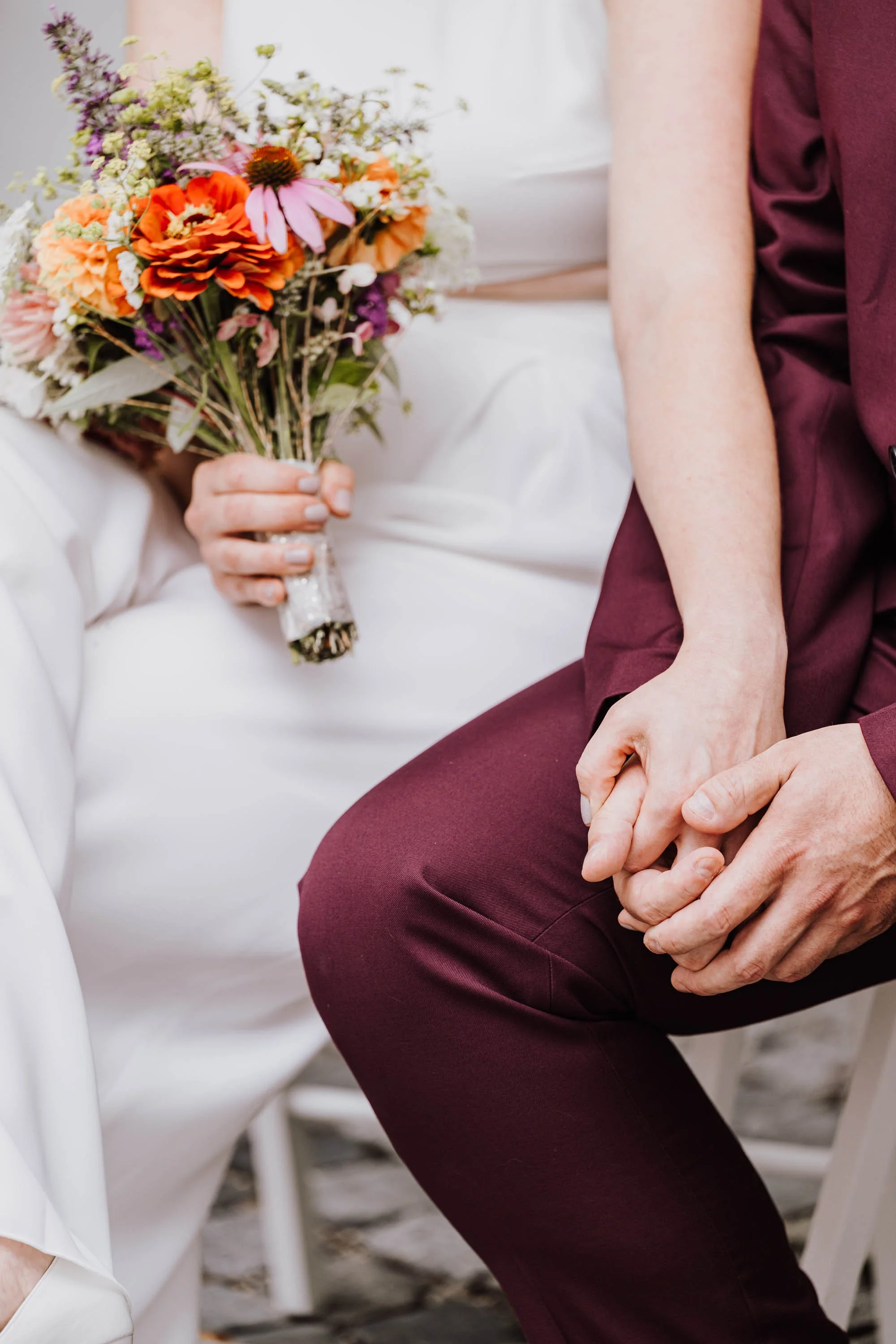 Nahaufnahme eines Paares bei einer Hochzeit, das sich an den Händen hält. Die Frau hält einen bunten Blumenstrauß in der Hand, während sie nebeneinander sitzen.