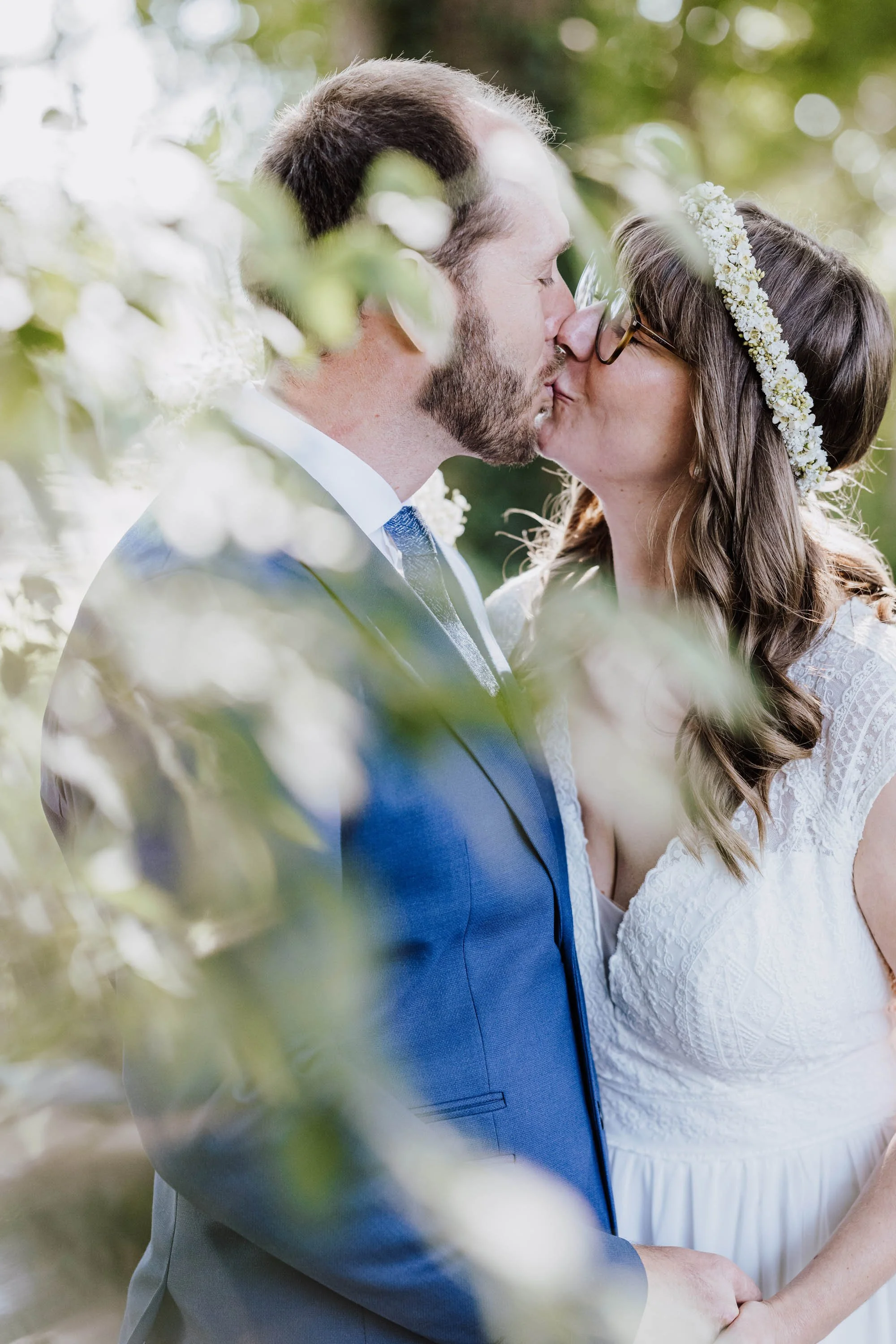 Braut und Bräutigam küssen sich im Freien, umgeben von grünen Blättern und Sonnenlicht.
