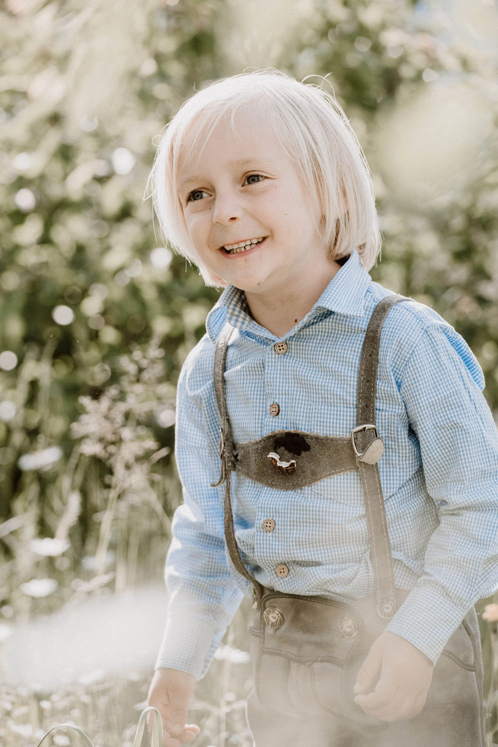 Ein Junge mit blonden Haaren, lächelnd, in einem blauen Hemd, steht im Freien, umgeben von Pflanzen.