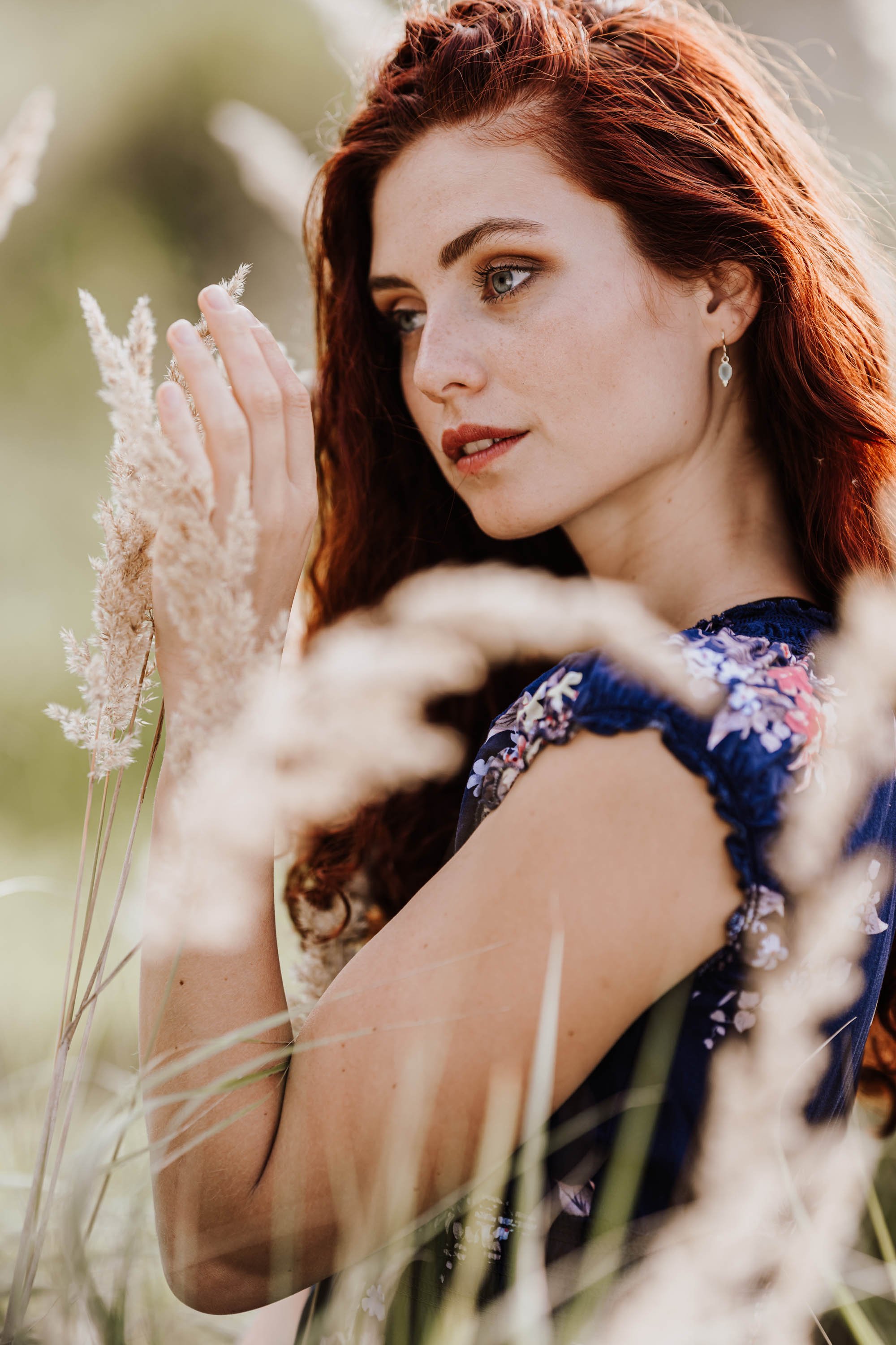 Junge Frau mit roten Haaren und Flecken im Gesicht, trägt ein dunkles Kleid mit floralem Muster, steht im Freien zwischen Gräsern und schaut nachdenklich zur Seite.