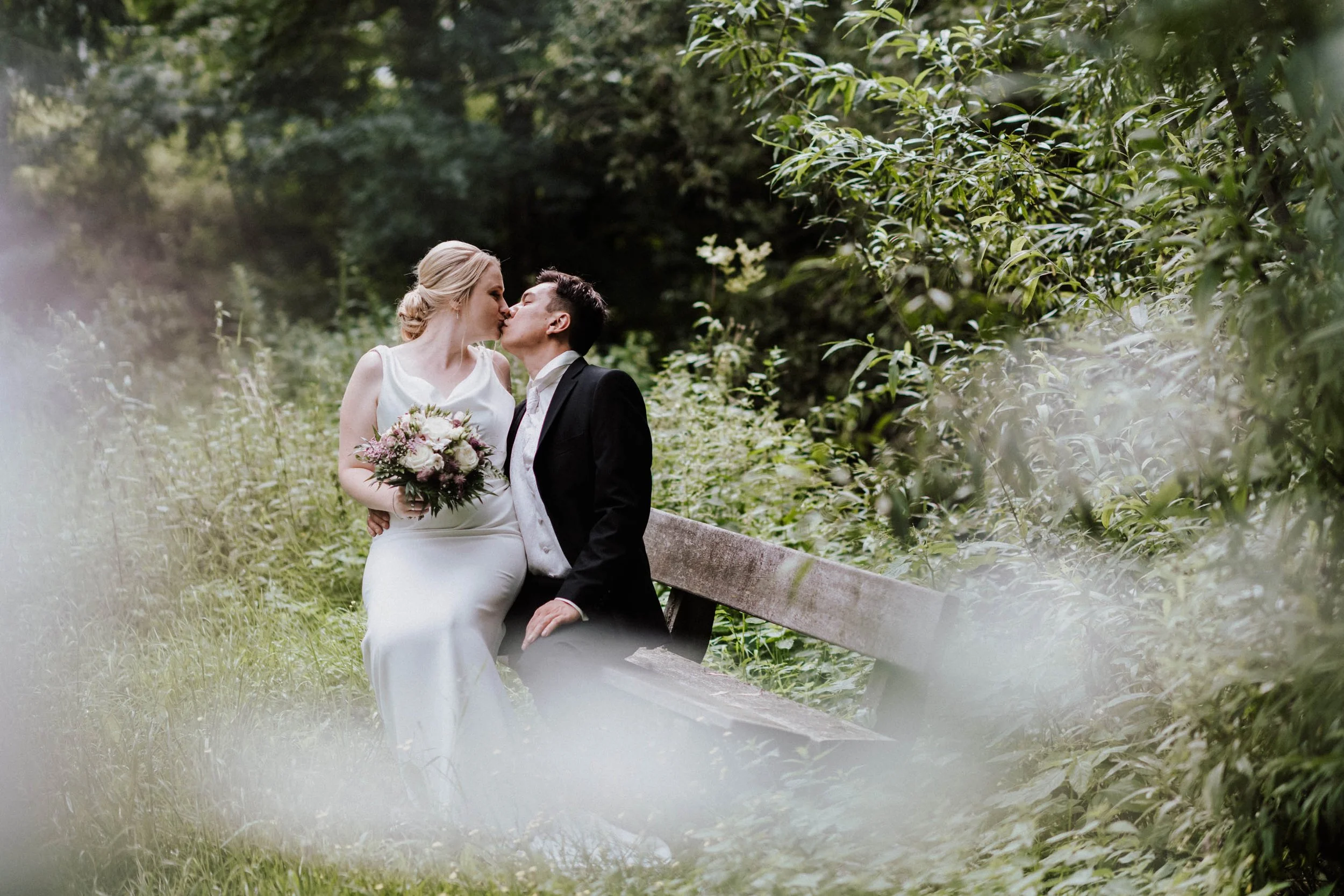 Ein Brautpaar sitzt auf einer Parkbank in einem Wald und küsst sich, die Braut hält einen Blumenstrauß.