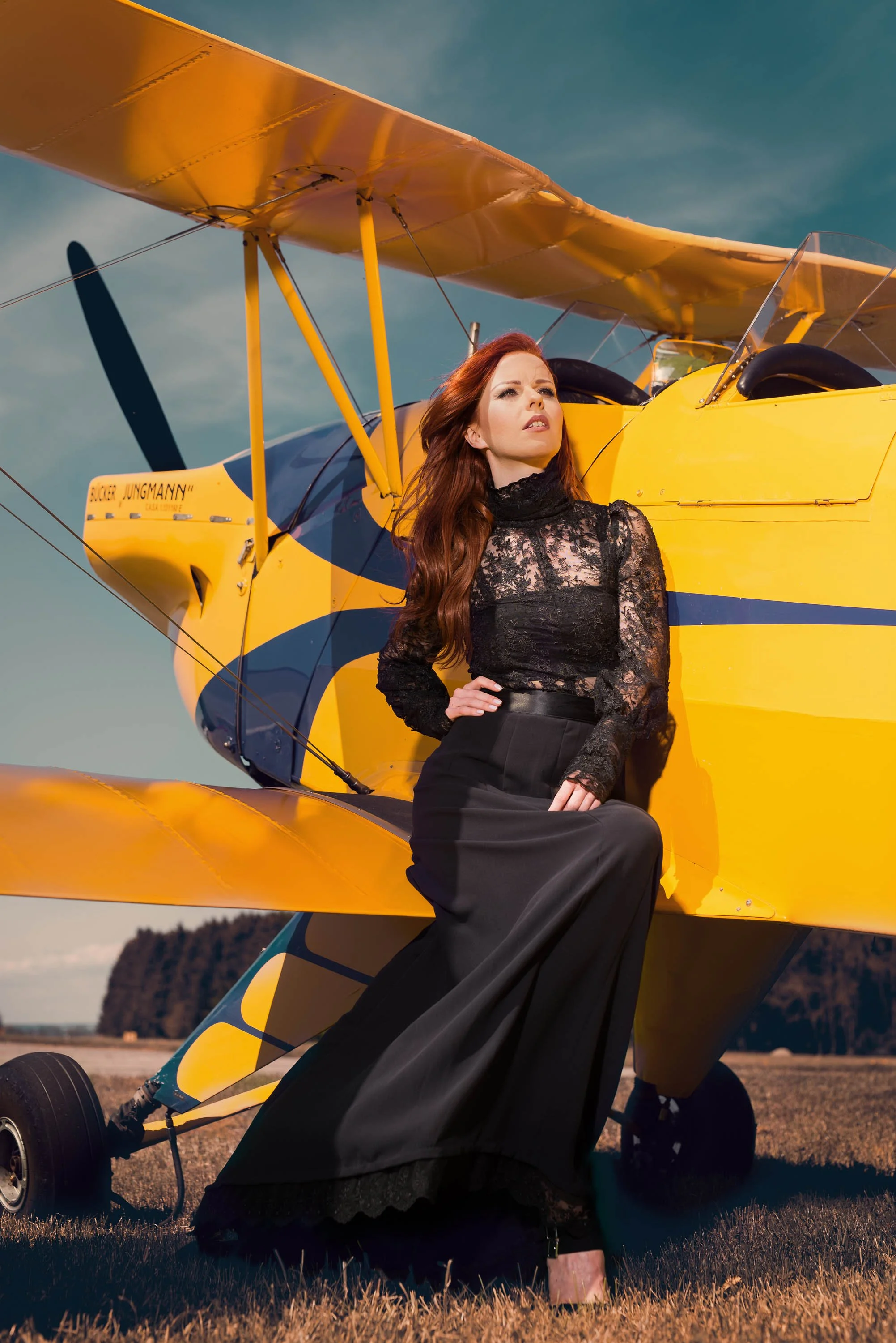Eine Frau in einem schwarzen Dirndl mit einer schwarzen Spitzenbluse sitzt auf dem Flügel eines gelben Flugzeugs, während sie in die Ferne blickt, auf einem Feld bei Sonnenuntergang.