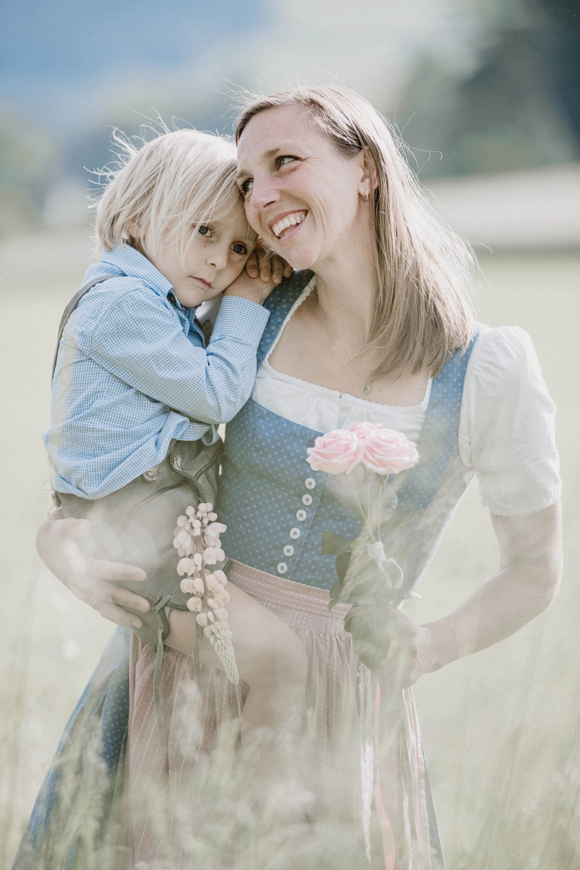 Eine Frau in traditioneller bayerischer Tracht trägt einen kleinen Jungen auf dem Arm. Beide halten einen Blumenstrauß. Sie stehen in einer Wiese mit einer bewölkten Himmel im Hintergrund. Die Frau lächelt fröhlich, während der Junge nachdenklich sch