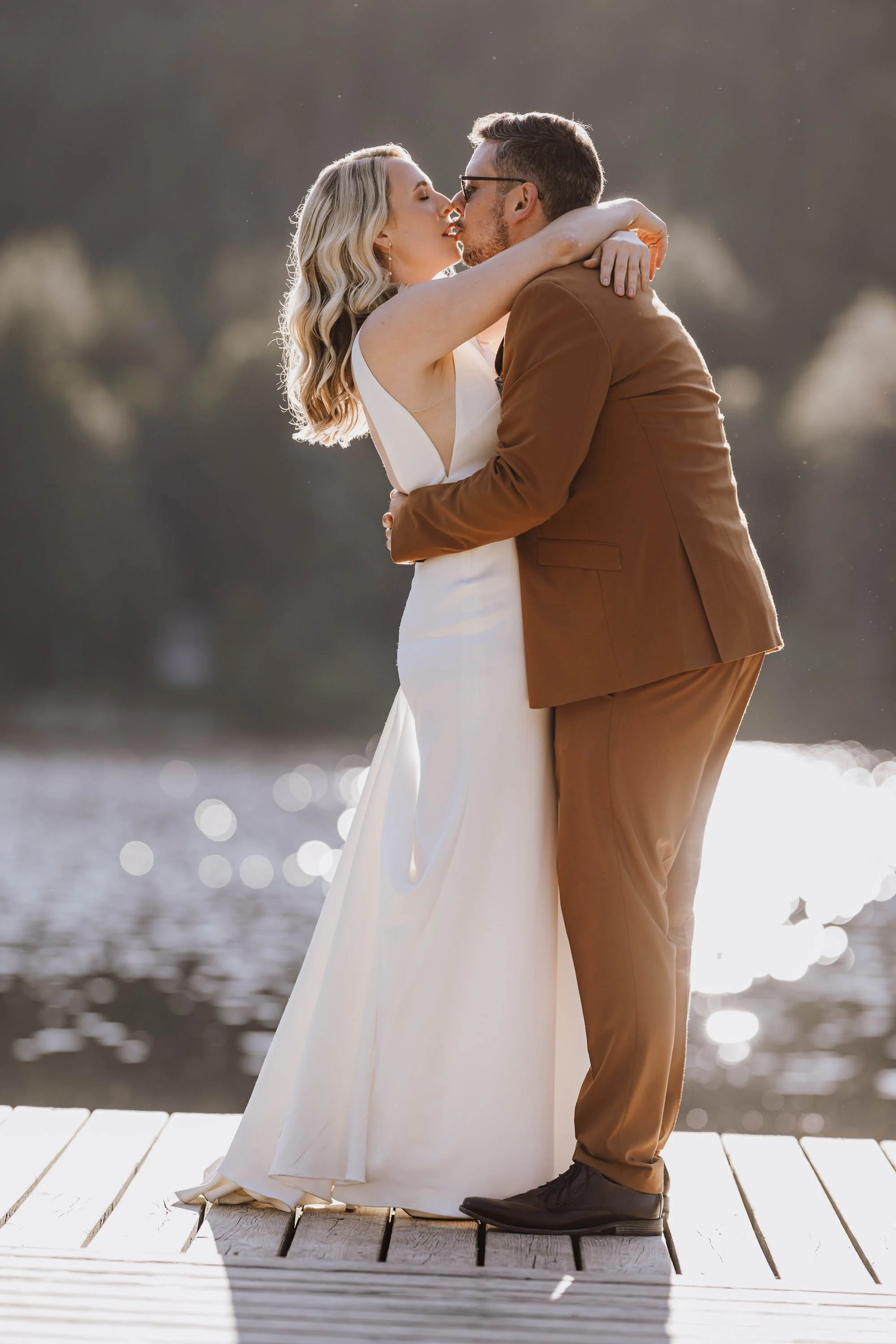 Ein Paar küsst sich liebevoll am Wasser bei Sonnenuntergang, auf einem Holzsteg, im Hochzeitsoutfit, im Freien.