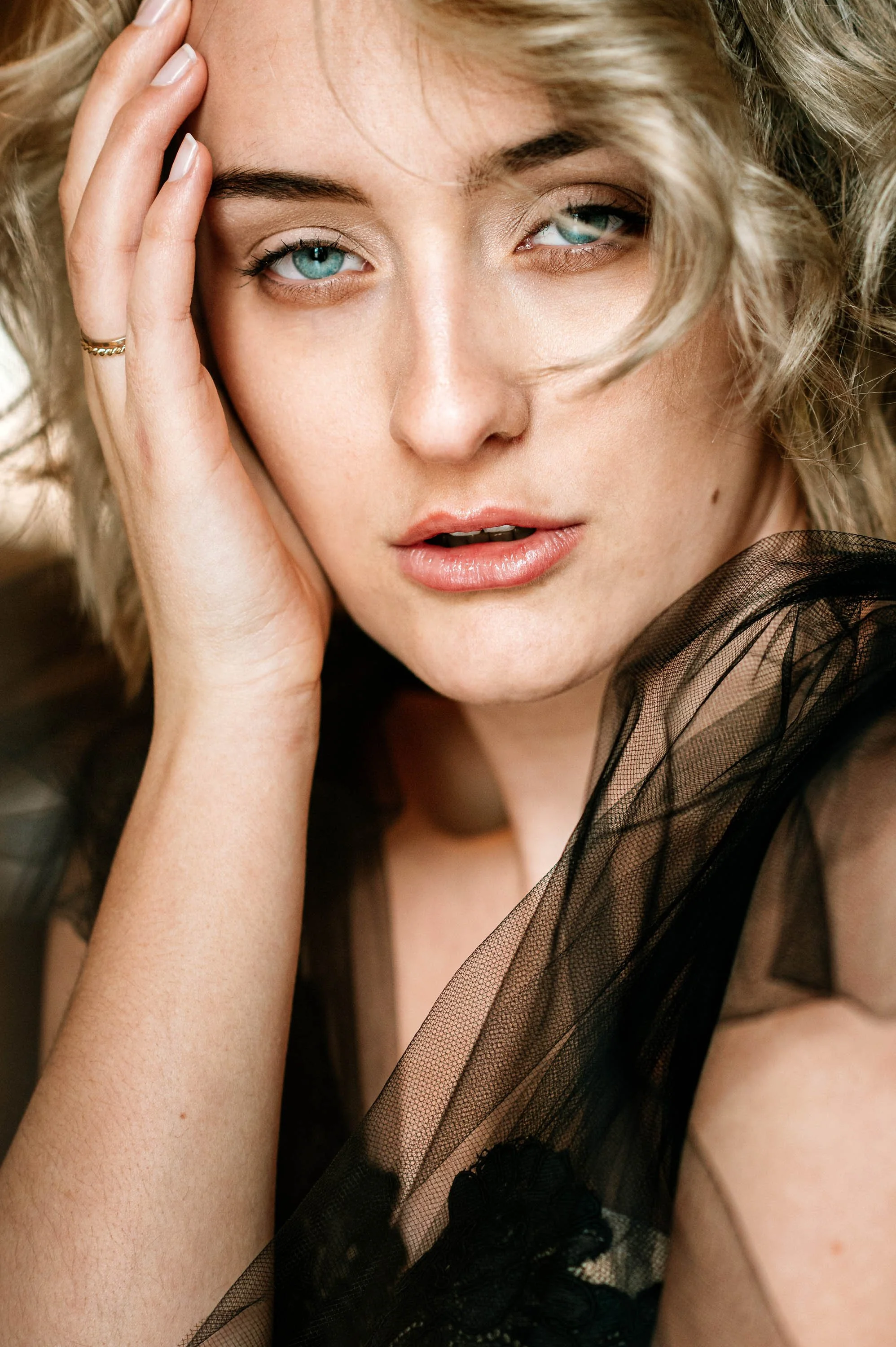 Nahaufnahme einer jungen Frau mit blonden, lockigen Haaren und blauen Augen, die ihre Hand an die Stirn legt und in die Kamera blickt, getragen in einem schwarzen, transparenten Spitzenkleid.