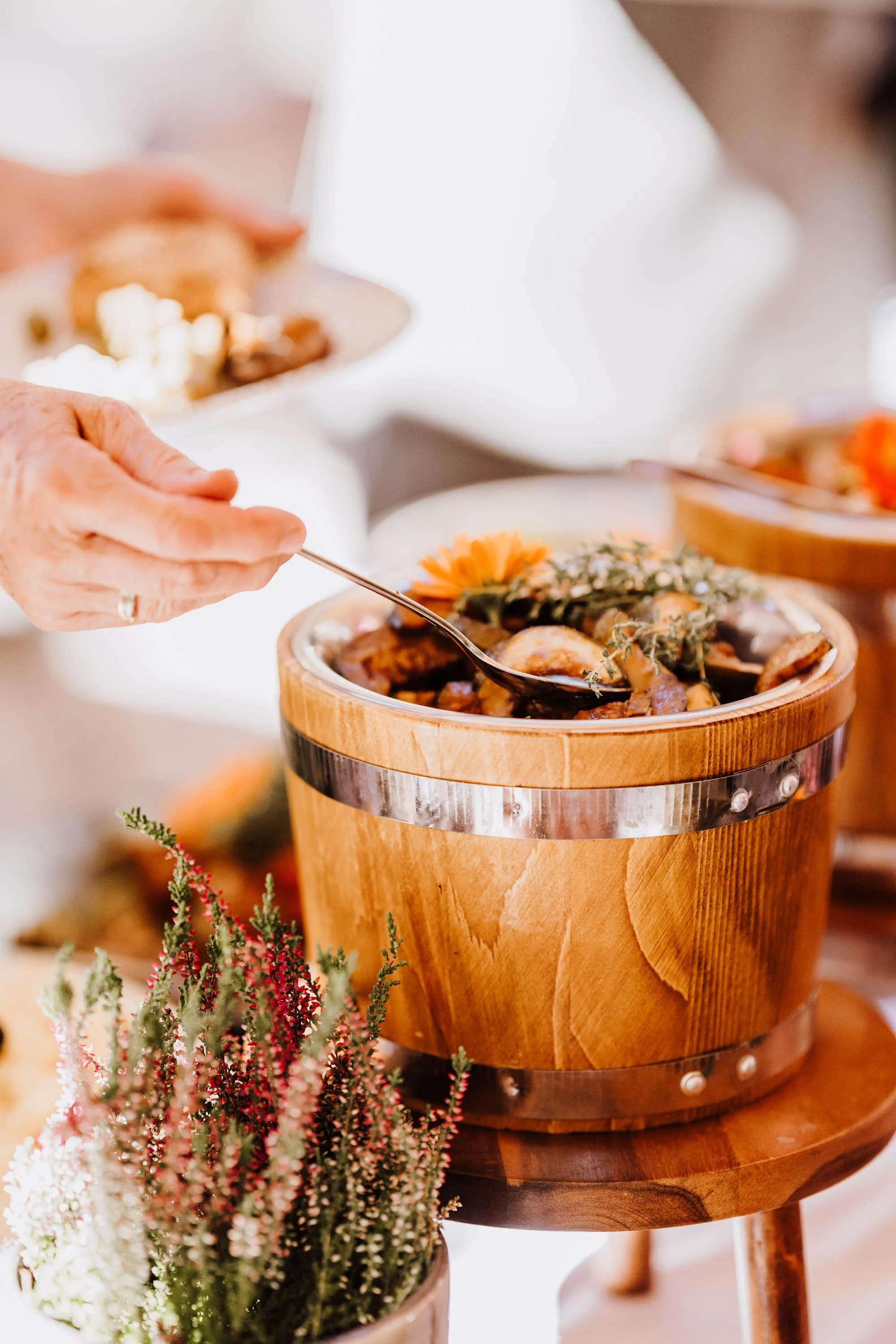 Nahaufnahme einer Person, die mit einem Löffel eine Schüssel mit Eintopf füllt. Die Schüssel besteht aus Holz und ist dekoriert mit einer kleinen Blume und Kräutern, auf einem kleinen Hocker. Im Vordergrund befindet sich eine Topfpflanze.