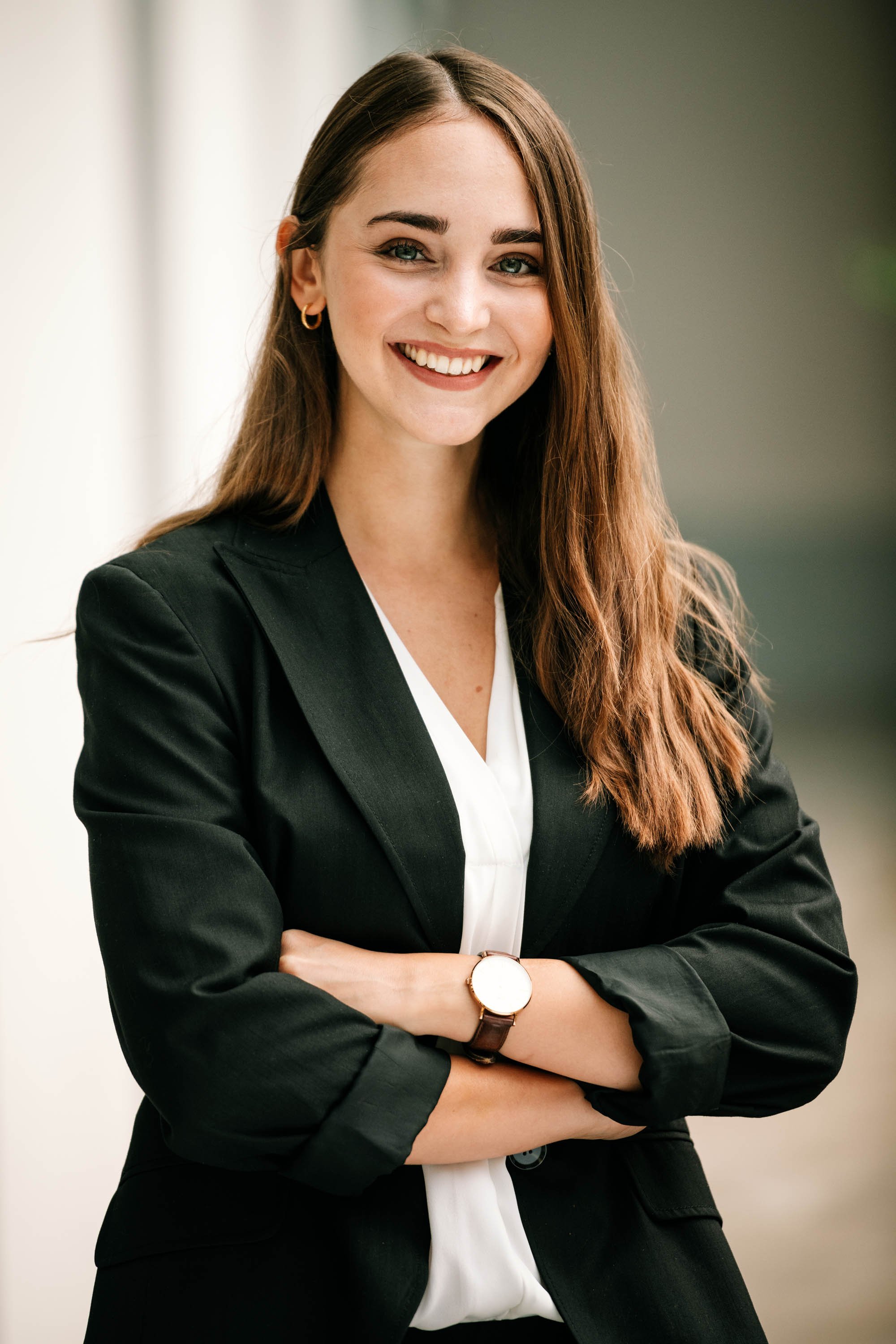 Ein lächelnder junges Frau mit braunen Haaren, trägt eine schwarze Jacke und eine weißen Bluse, steht mit verschränkten Armen vor einem neutralen Hintergrund.