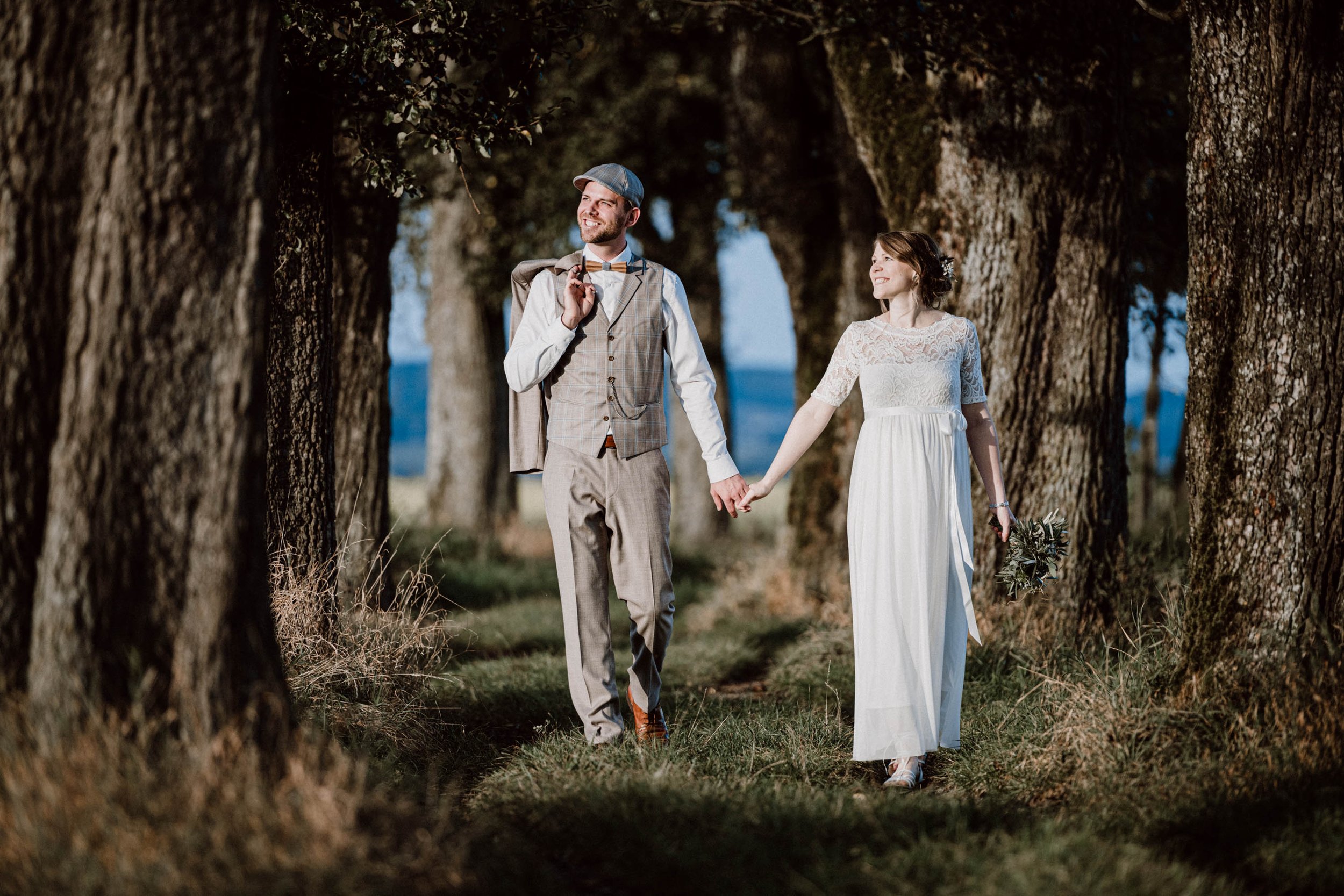 Ein frisch verheiratetes Paar spaziert Hand in Hand durch einen Baumweg im Freien bei Sonnenuntergang. Die Frau trägt ein langesweißes Brautkleid und hält einen Blumenstrauß, während der Mann einen Anzug mit Weste, Krawatte und einem Hut trägt. Sie l