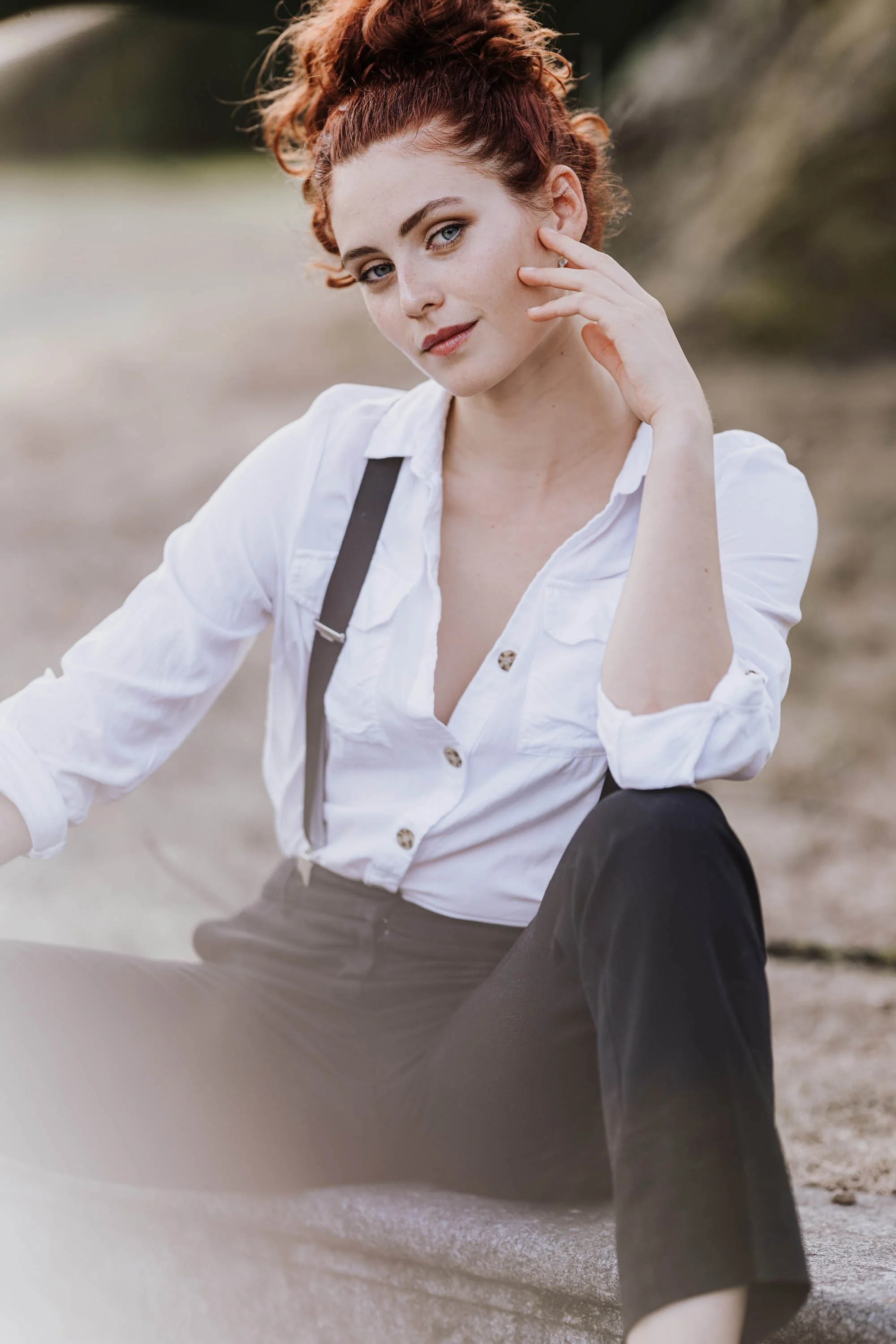 Junge Frau mit roten, lockigen Haaren, sitzend im Freien auf einem Felsen, trägt weiße Bluse und schwarze Hosen, schaut in die Kamera.