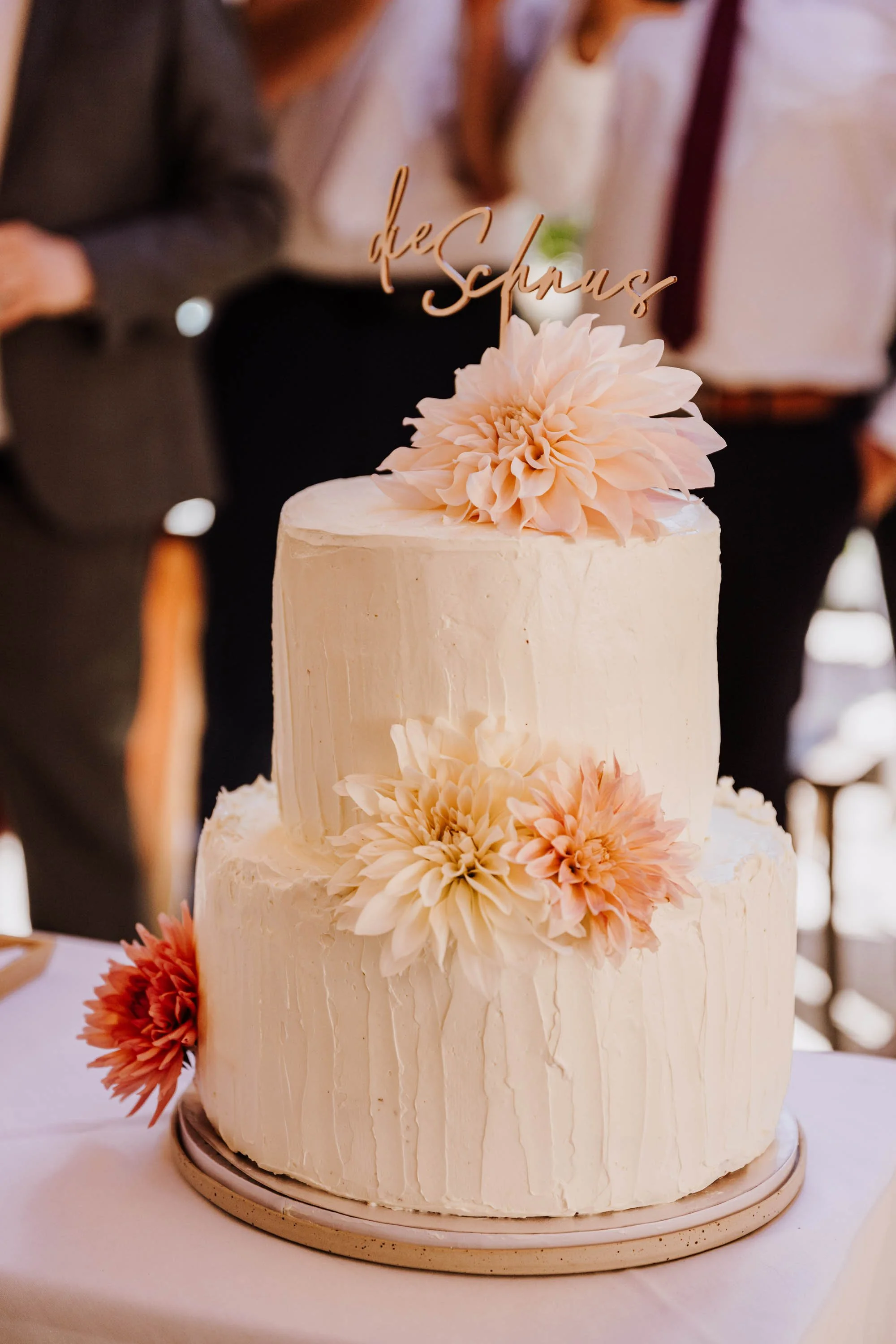 Zweistöckige weiße Hochzeitstorte mit Blüten und einem goldfarbenen Schriftzug 'die Schnus' auf der Spitze bei einer Hochzeitsfeier.