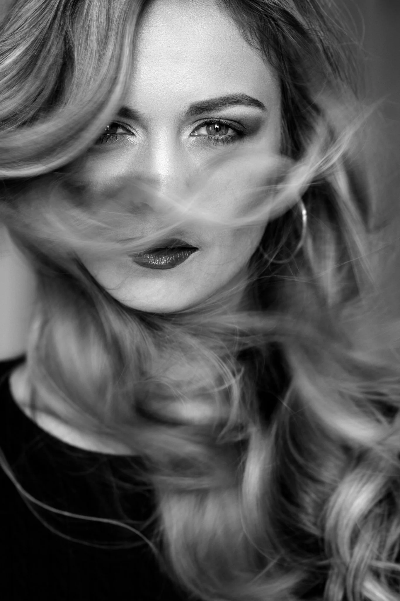 Schwarz-weiß-Bild eines Gesichts einer Frau mit langen, gewellten Haaren und intensiven Blick, teilweise von Haaren bedeckt.