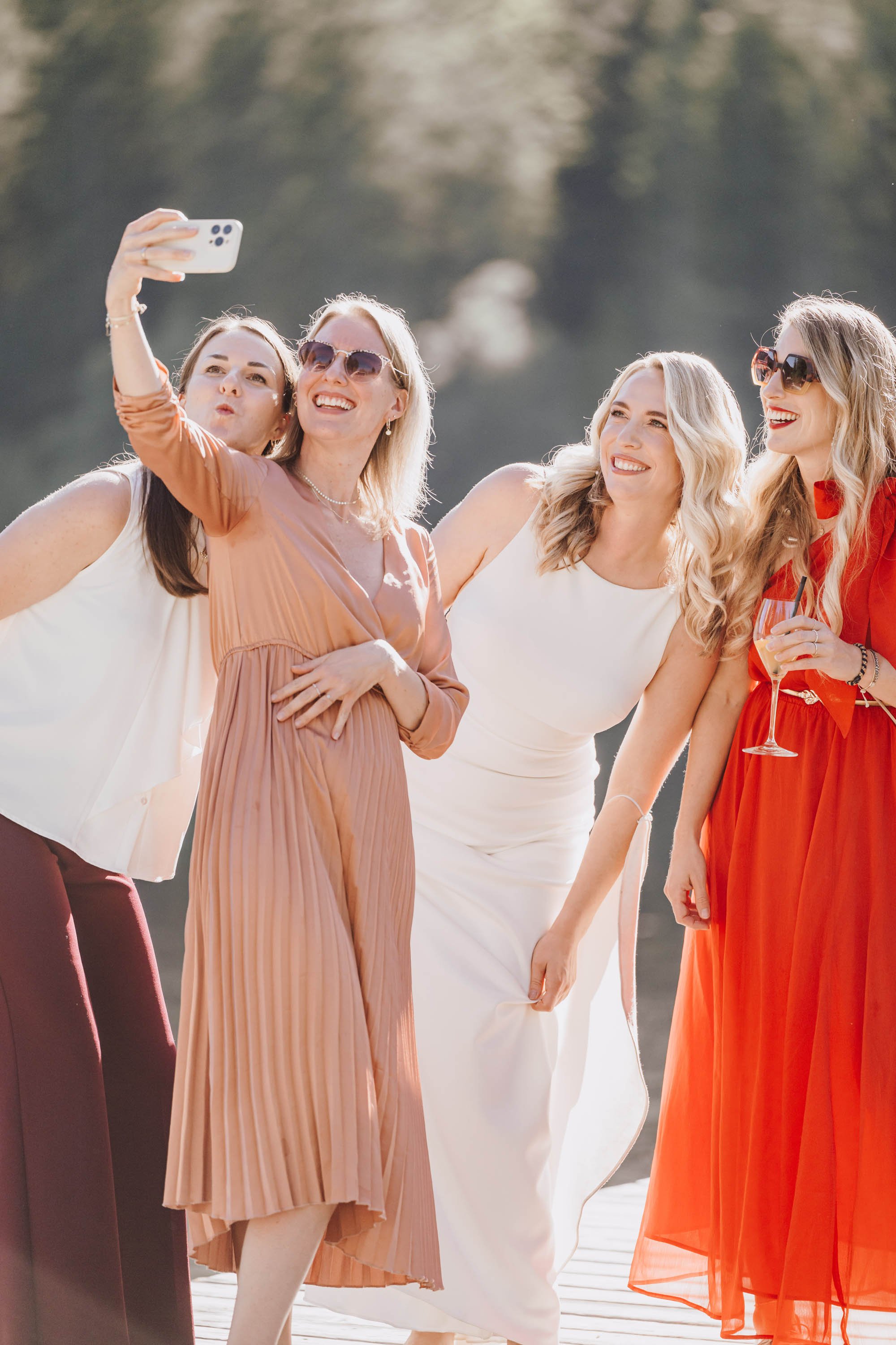 Fünf Frauen machen ein Selfie und lachen an einem sonnigen Tag draußen am Wasser.
