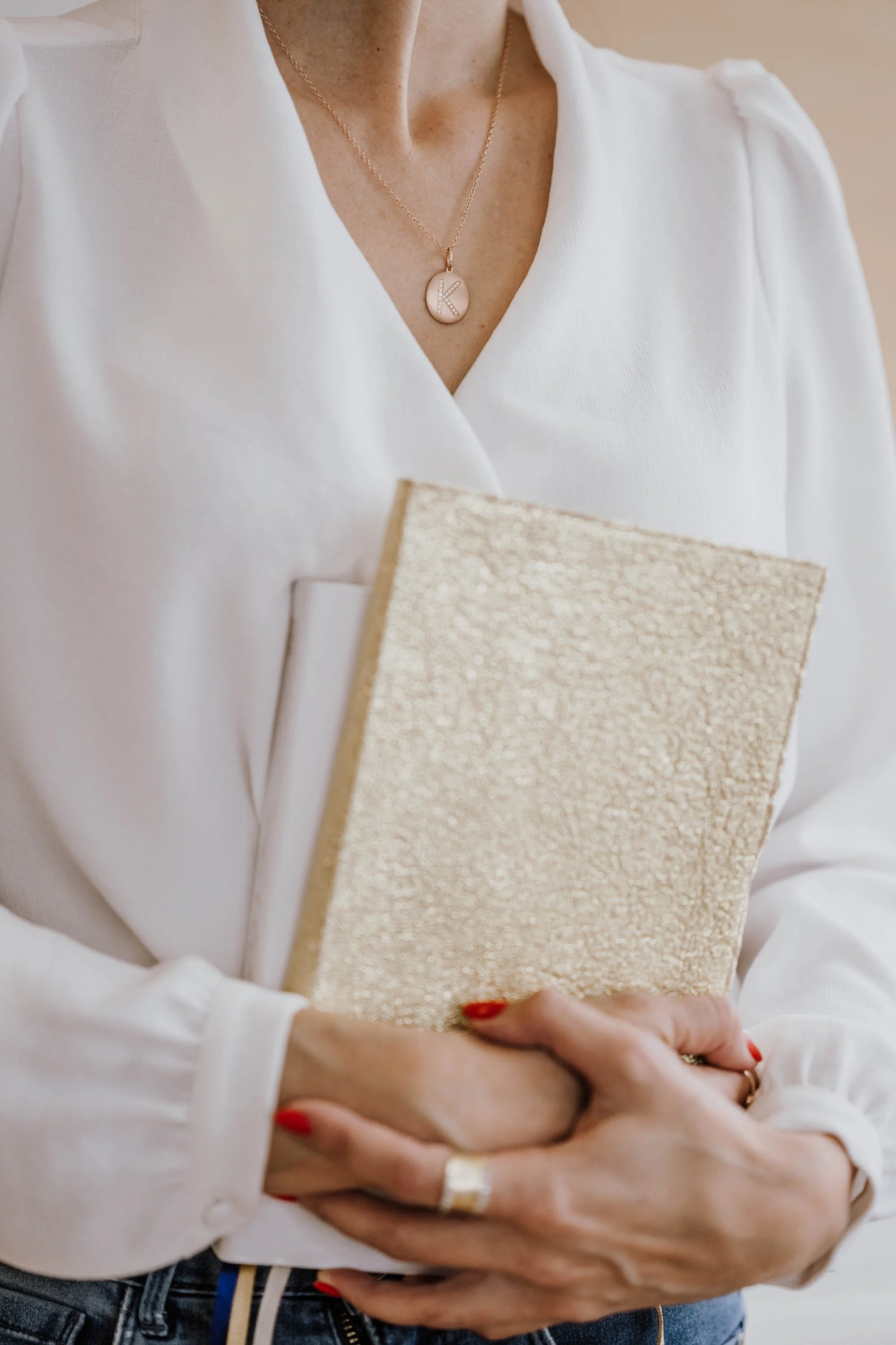 Nahaufnahme einer Frau, die ein beige Buch hält, trägt eine weiße Bluse, goldene Halskette mit einem Medaillon mit dem Buchstaben 'K', und rotem Nagellack.