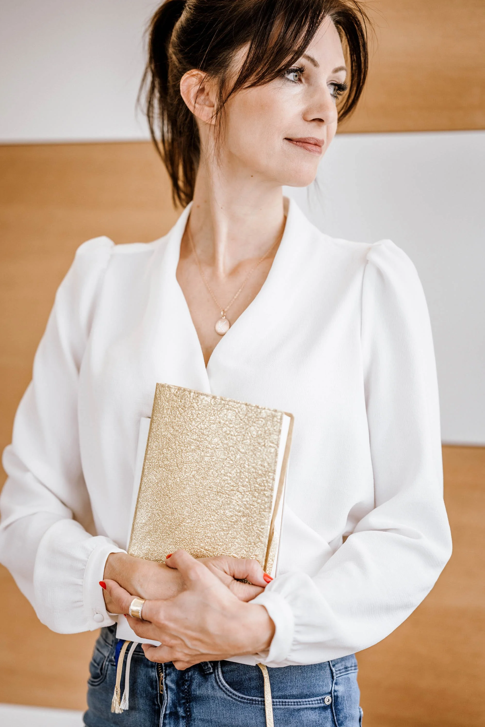 Eine Frau im weißen Hemd hält ein goldenes Notizbuch vor sich, steht vor einer Holzwand, trägt eine Halskette und einen Ring.